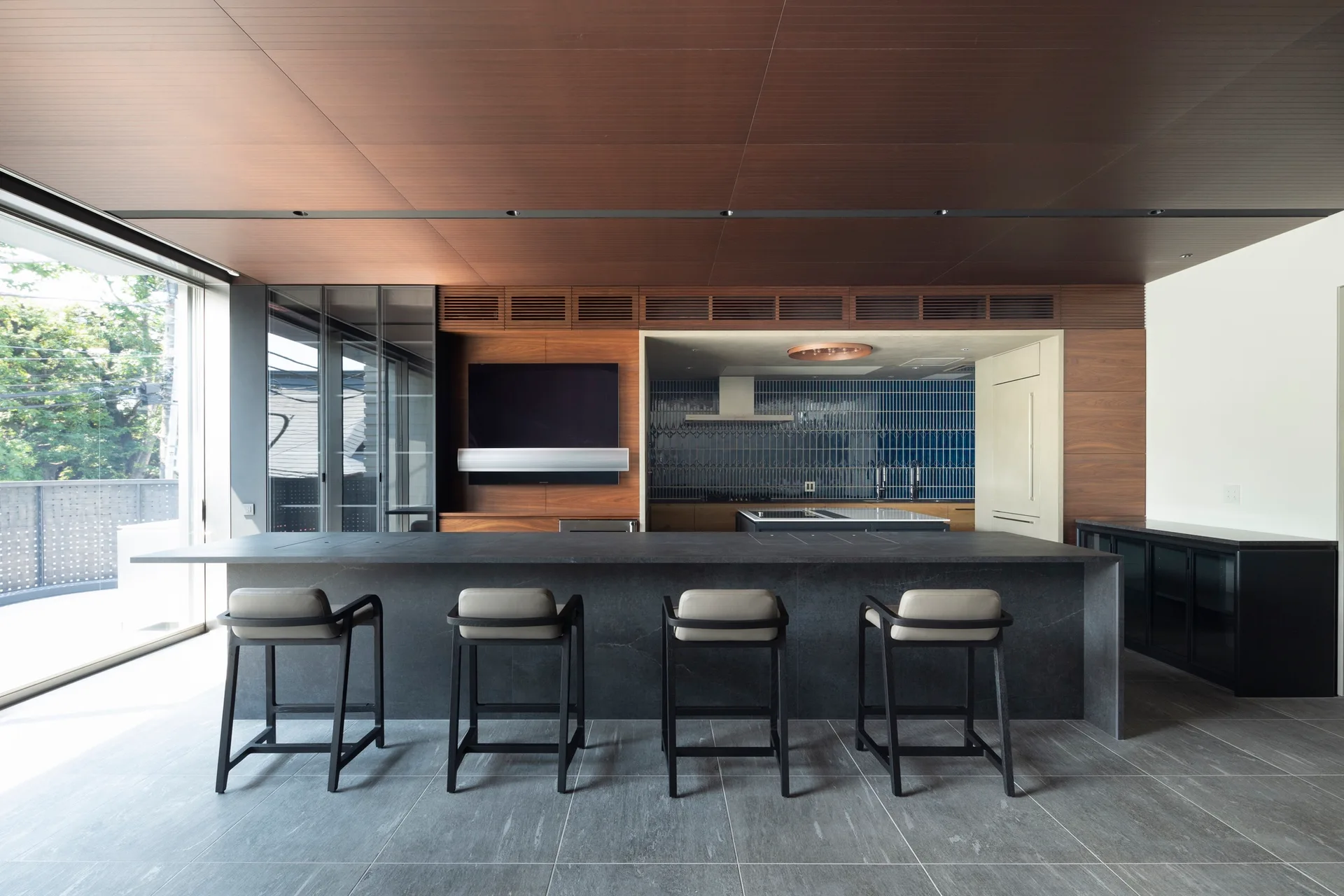 A modern kitchen features a large dark stone island with four bar stools, a wood-paneled wall and ceiling, and a blue vertical tile backsplash.