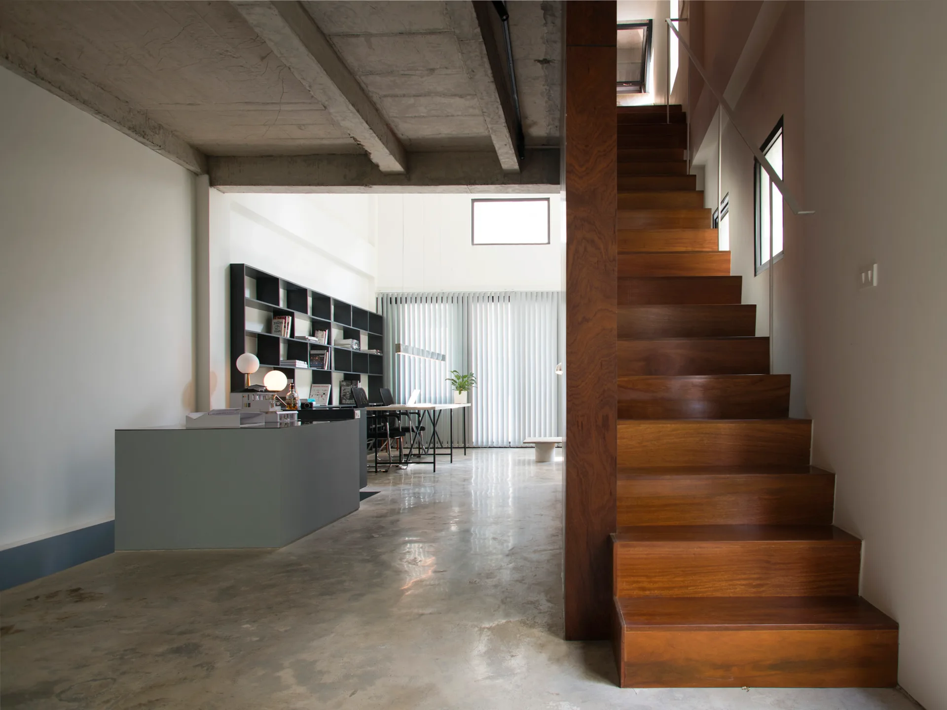 A modern home office features a wooden staircase in the foreground, polished concrete floors, and a desk next to a black bookshelf.