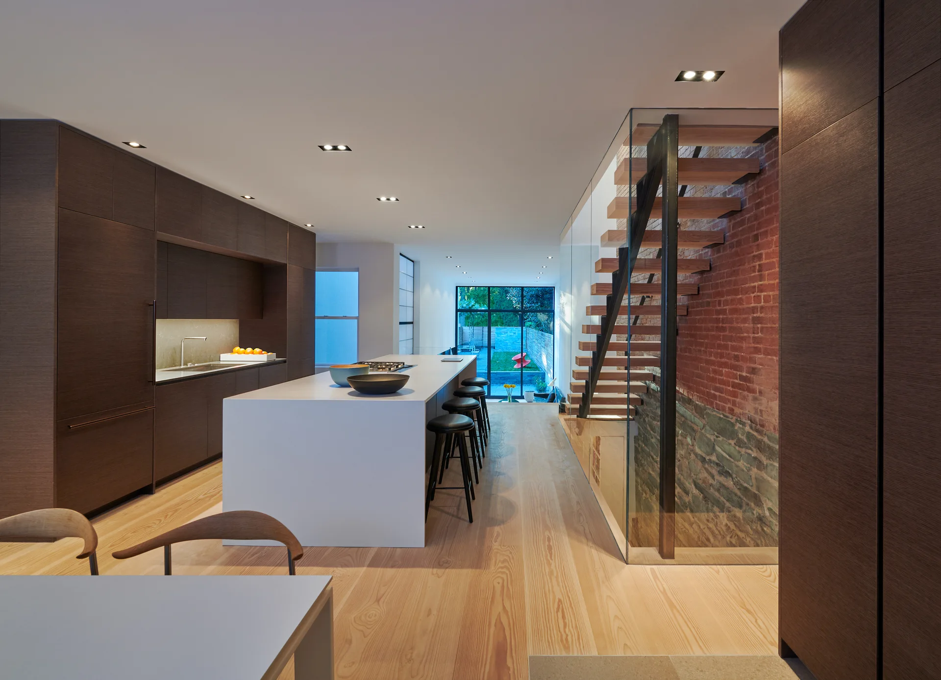 A modern kitchen features a long white island, dark wood cabinets, and a floating wood staircase with a glass railing against an exposed brick wall.
