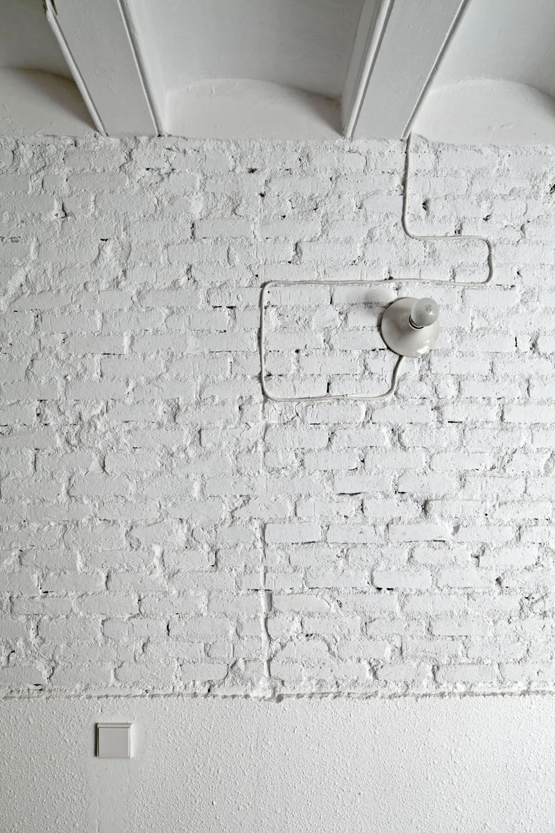 A white painted brick wall features a surface-mounted white cord routed in a geometric path to a simple white light fixture.