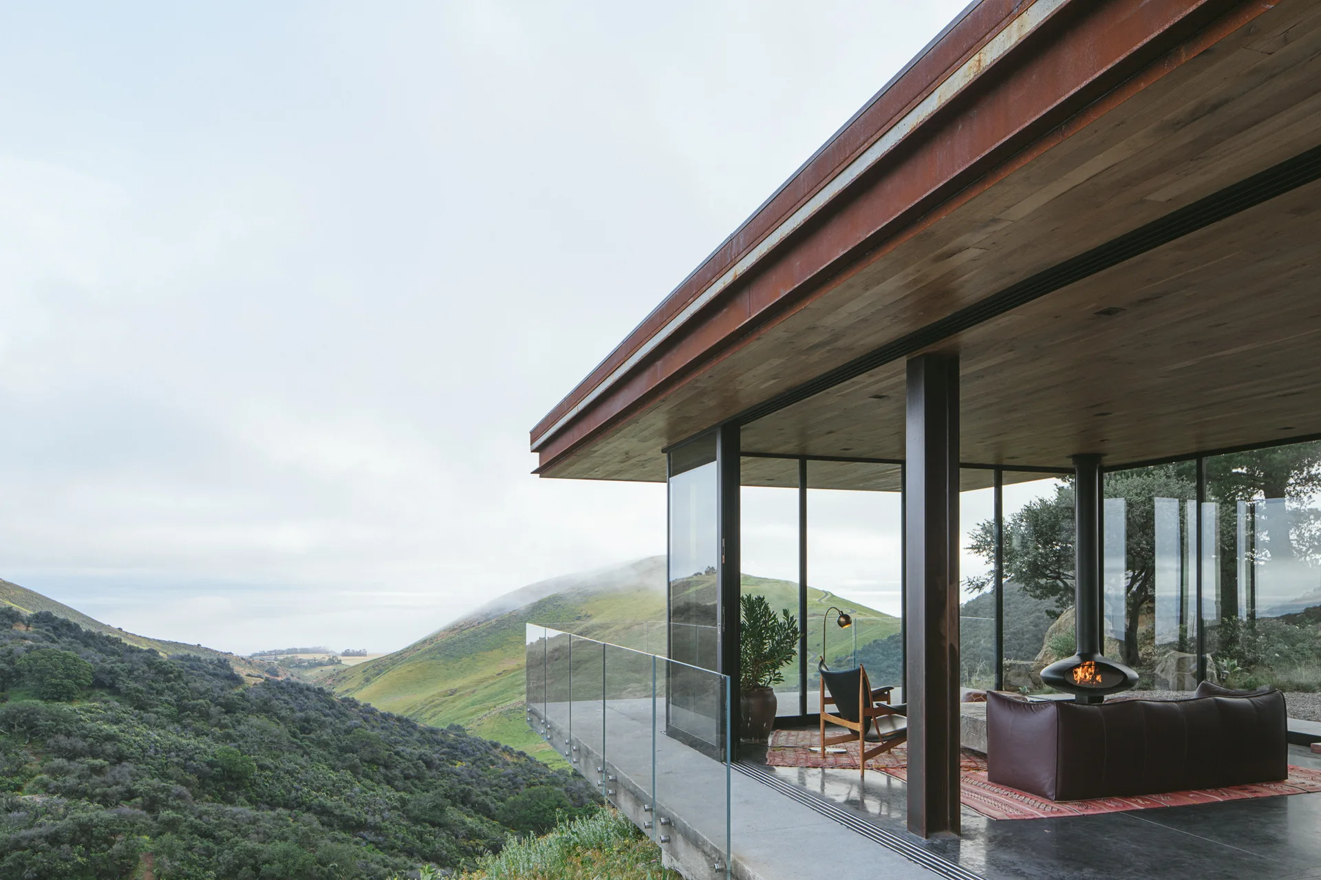 A modern living room with glass walls, a suspended fireplace, and a leather sofa overlooks a green hilly landscape from an outdoor deck.
