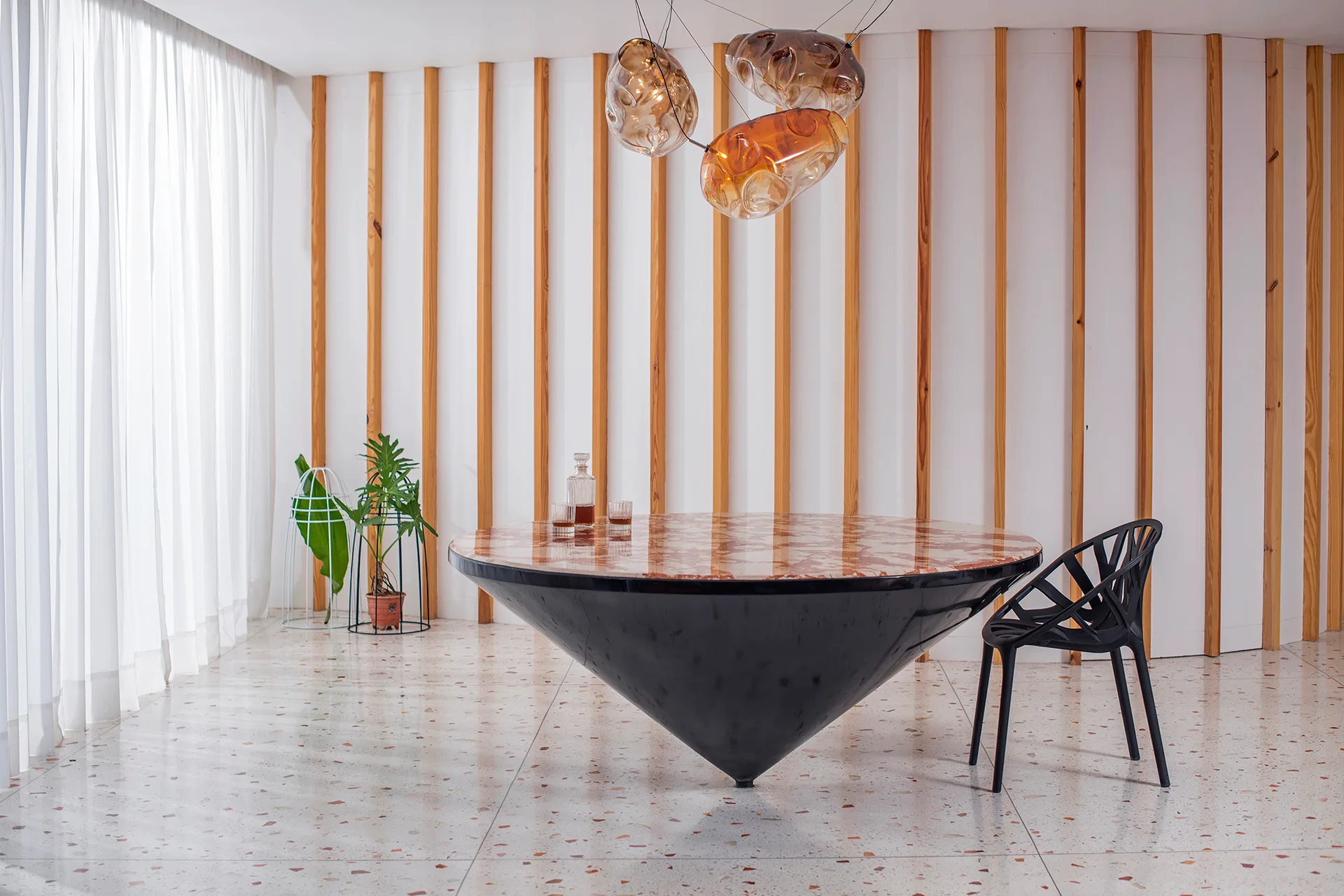 A modern dining room features a round black table with a conical base, a sculptural chair, terrazzo flooring, and a vertical wood slat wall.