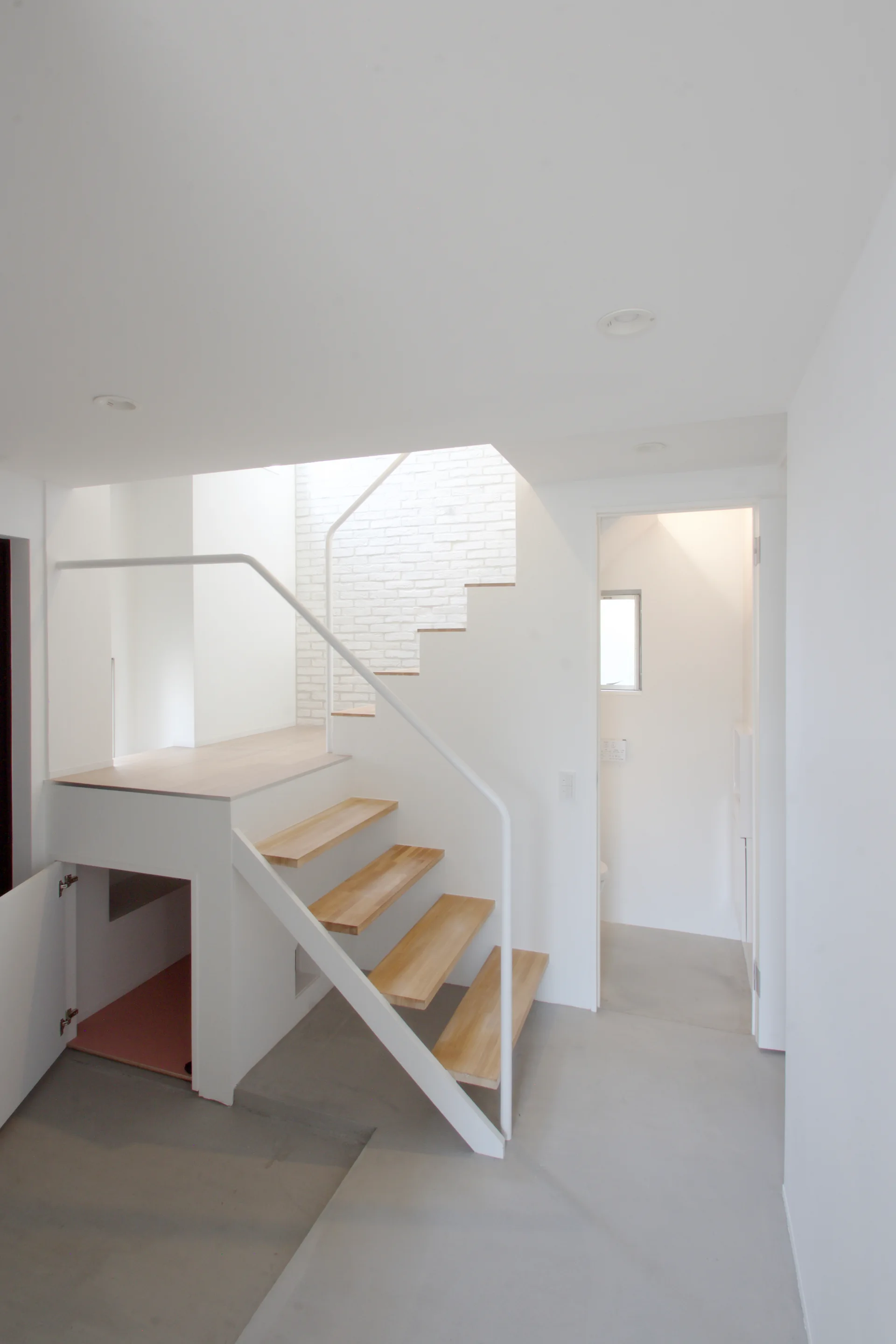 A minimalist white hallway featuring a floating staircase with light wood treads, a white metal handrail, and built-in storage underneath.