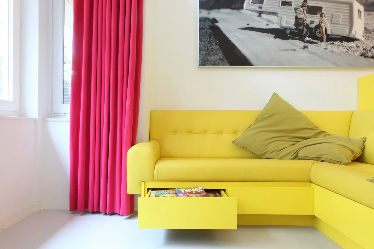 A bright yellow sectional sofa with an open under-seat storage drawer sits next to vibrant pink curtains and under framed wall art.