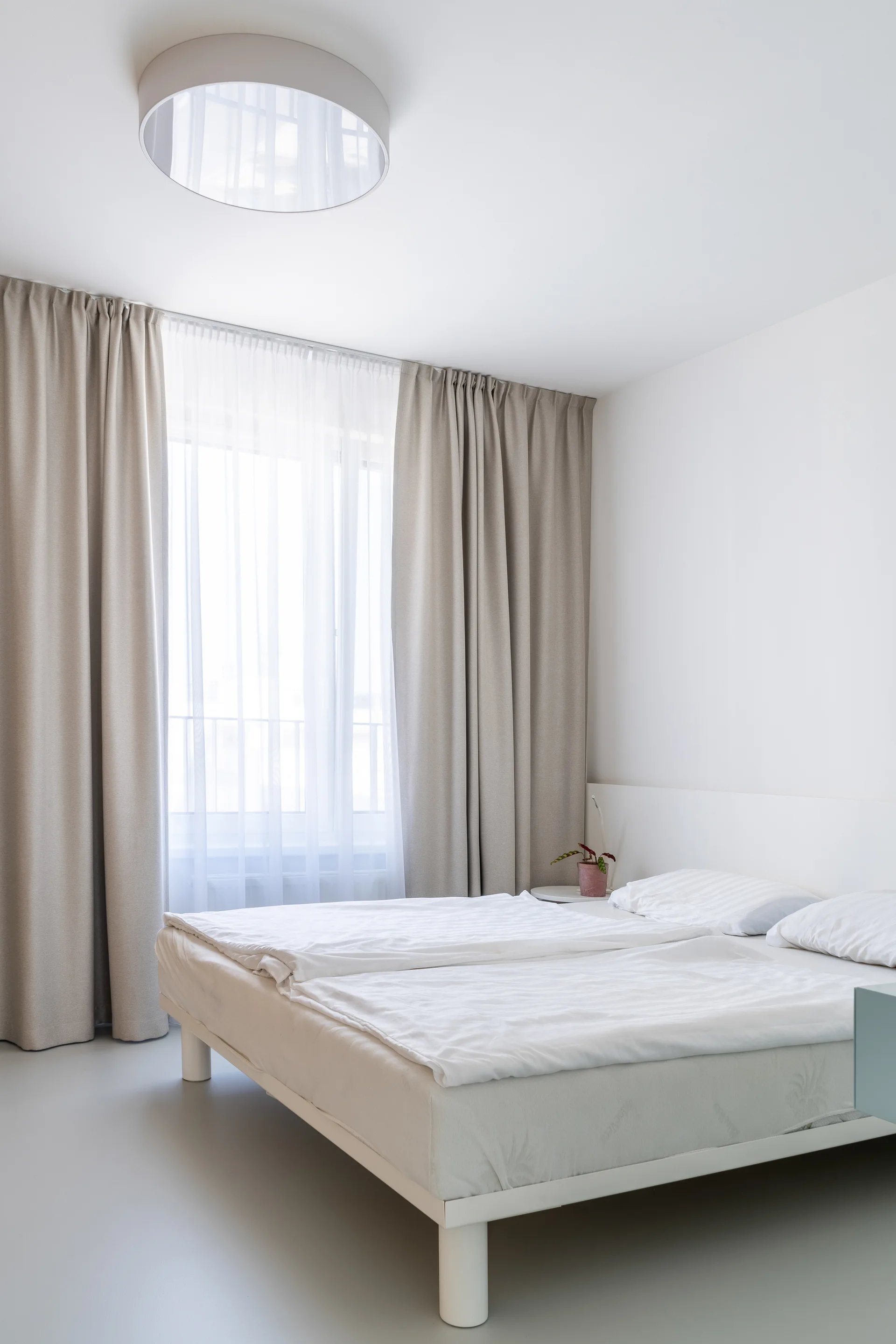 A minimalist white bedroom features a bed with white linens, floor-length beige curtains framing a large window, and a circular ceiling light.