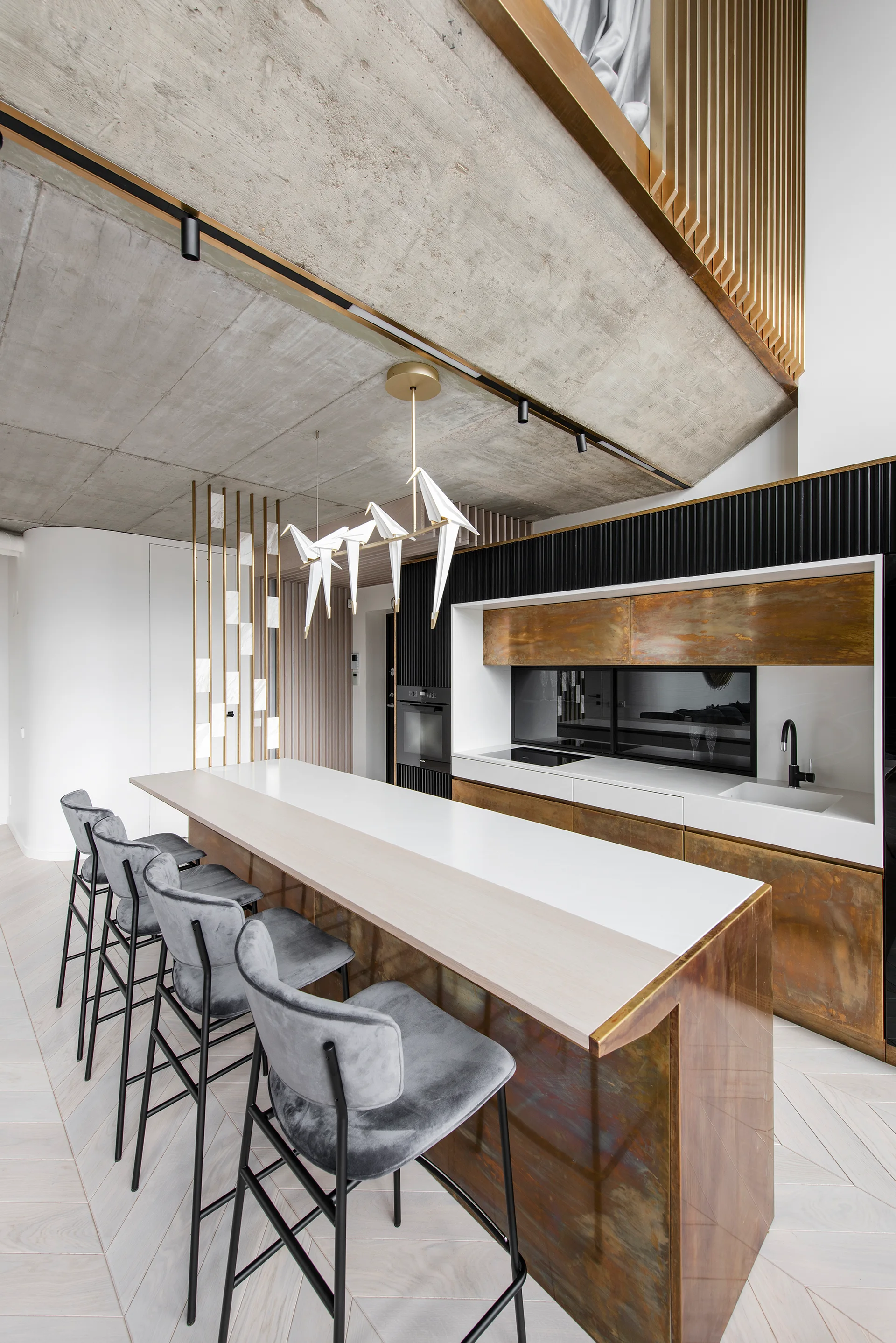 A modern kitchen features a bronze-faced island with grey velvet bar stools under an exposed concrete ceiling and bird-shaped pendant lights.