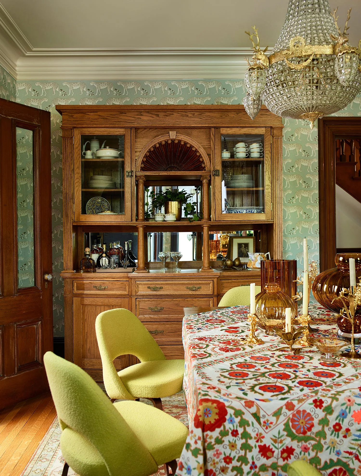 A dining room features a large wood china cabinet against cheetah-patterned wallpaper next to lime green chairs and a table with a floral tablecloth.