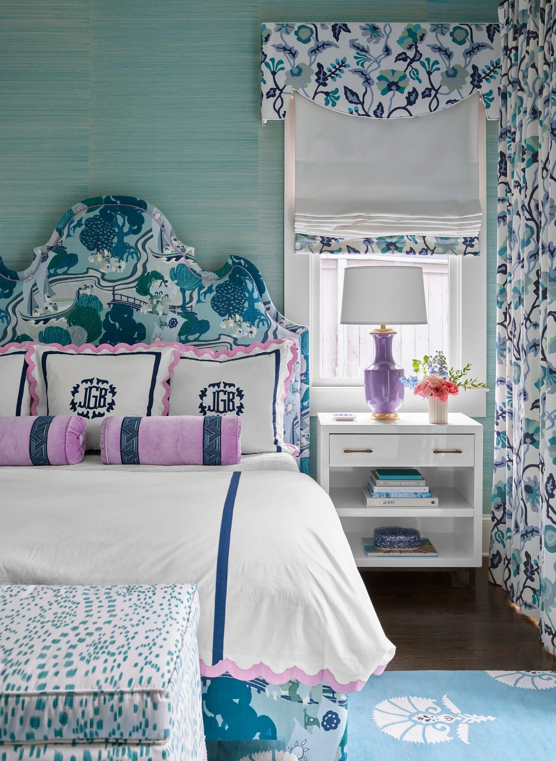 Bedroom with a patterned blue headboard, monogrammed shams with pink trim, teal grasscloth wallpaper, and a purple lamp on a white nightstand.