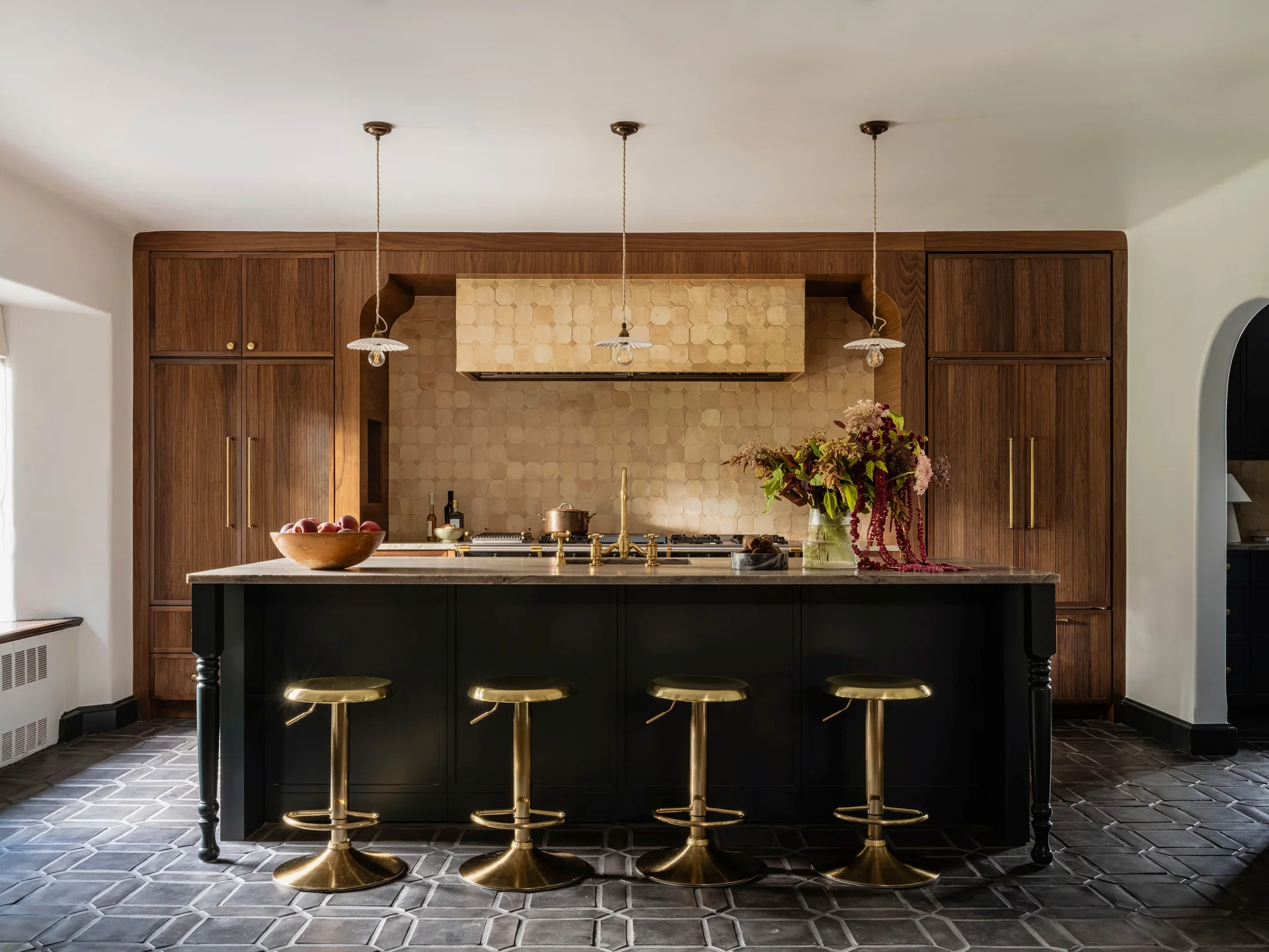 A dark kitchen island with brass stools sits in front of fluted wood cabinets and a hexagonal tile backsplash.