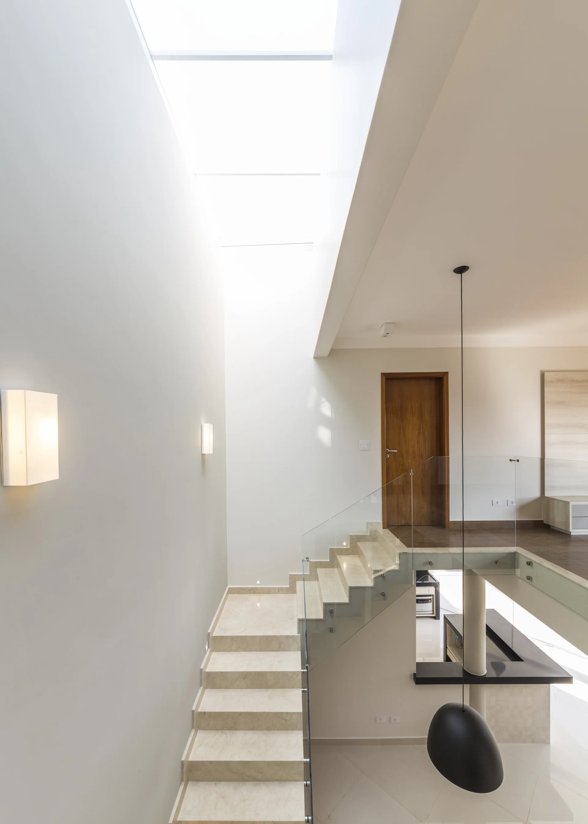 A minimalist staircase with light stone steps and glass railings rises toward a wooden door under a large overhead skylight.