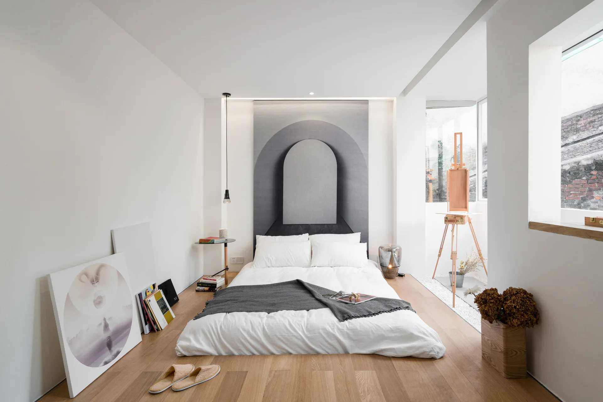 A minimalist bedroom featuring a low-profile bed, an archway graphic wall hanging, and a wooden artist easel next to a window.