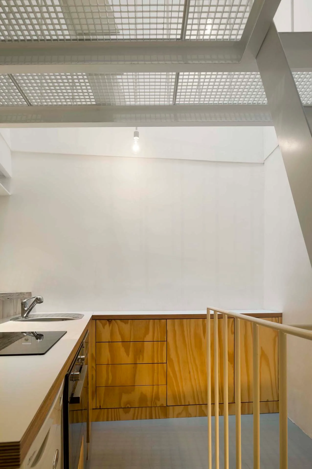 A minimalist kitchen features natural plywood cabinetry under a white metal grid ceiling structure.