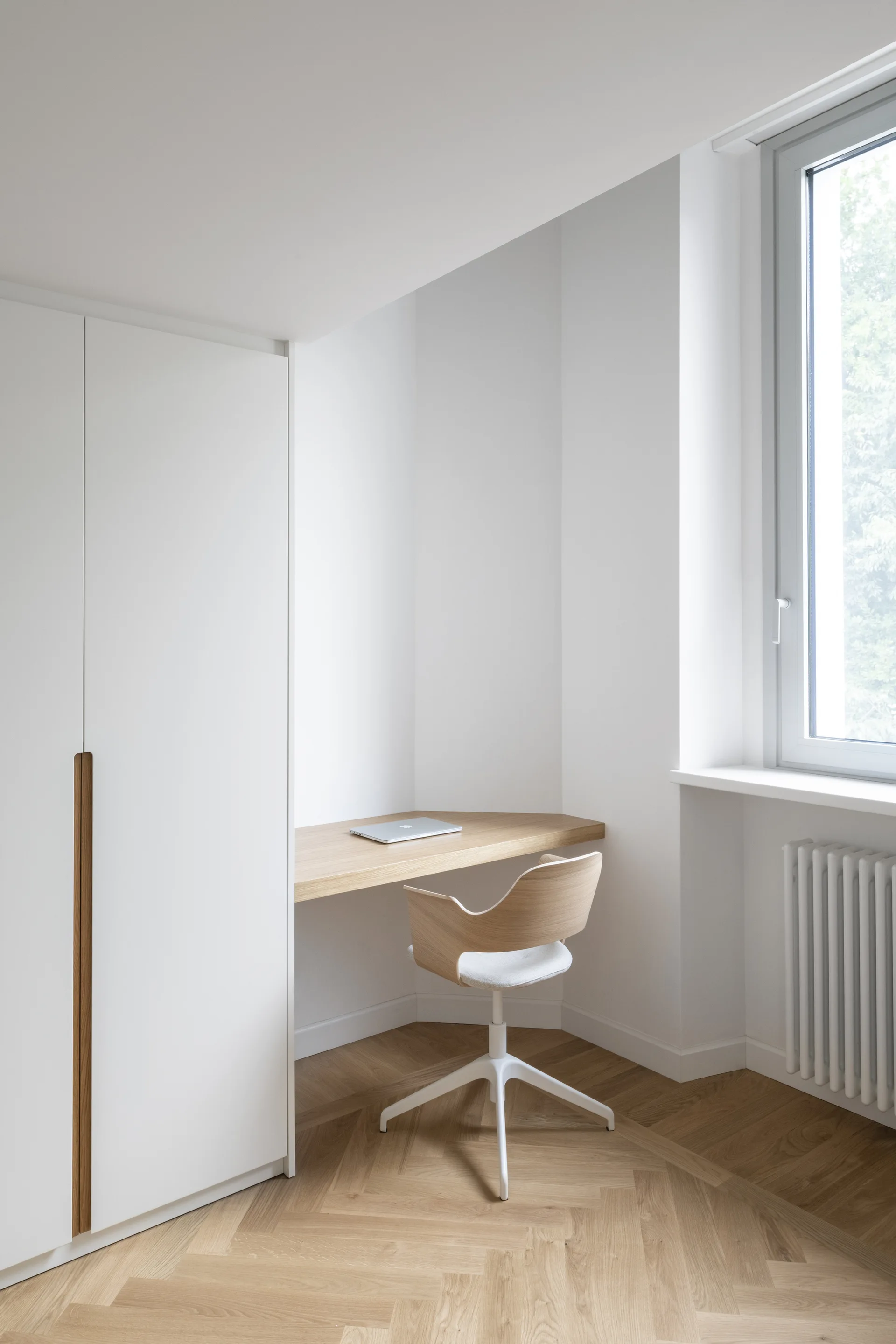 A minimalist home office corner features a built-in light wood desk, a matching curved swivel chair, white cabinetry, and herringbone wood floors.