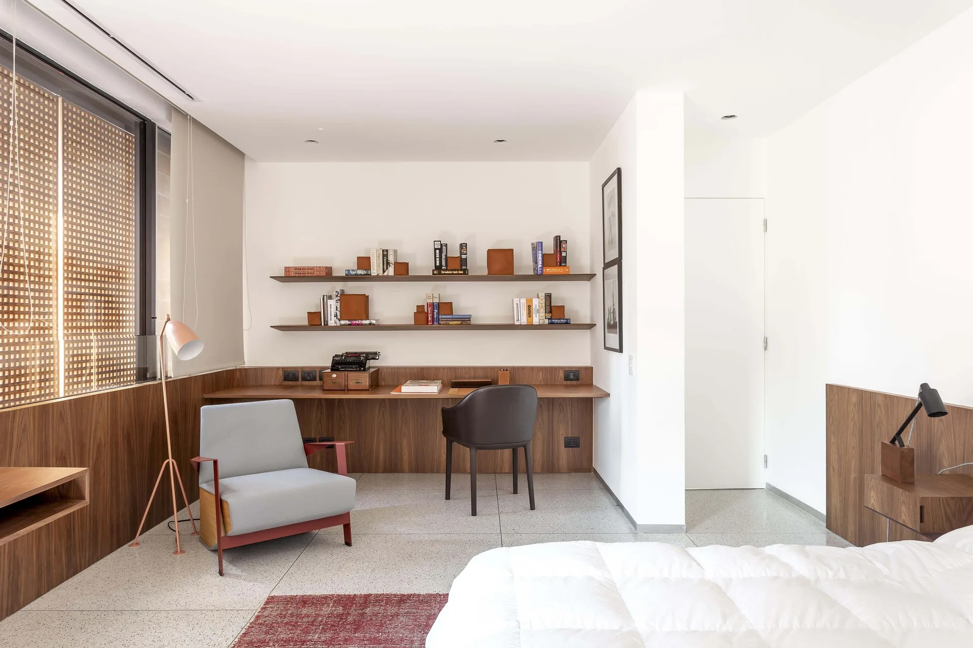A minimalist bedroom workspace features a built-in wooden desk, floating shelves with books, a grey armchair, and a terrazzo floor.