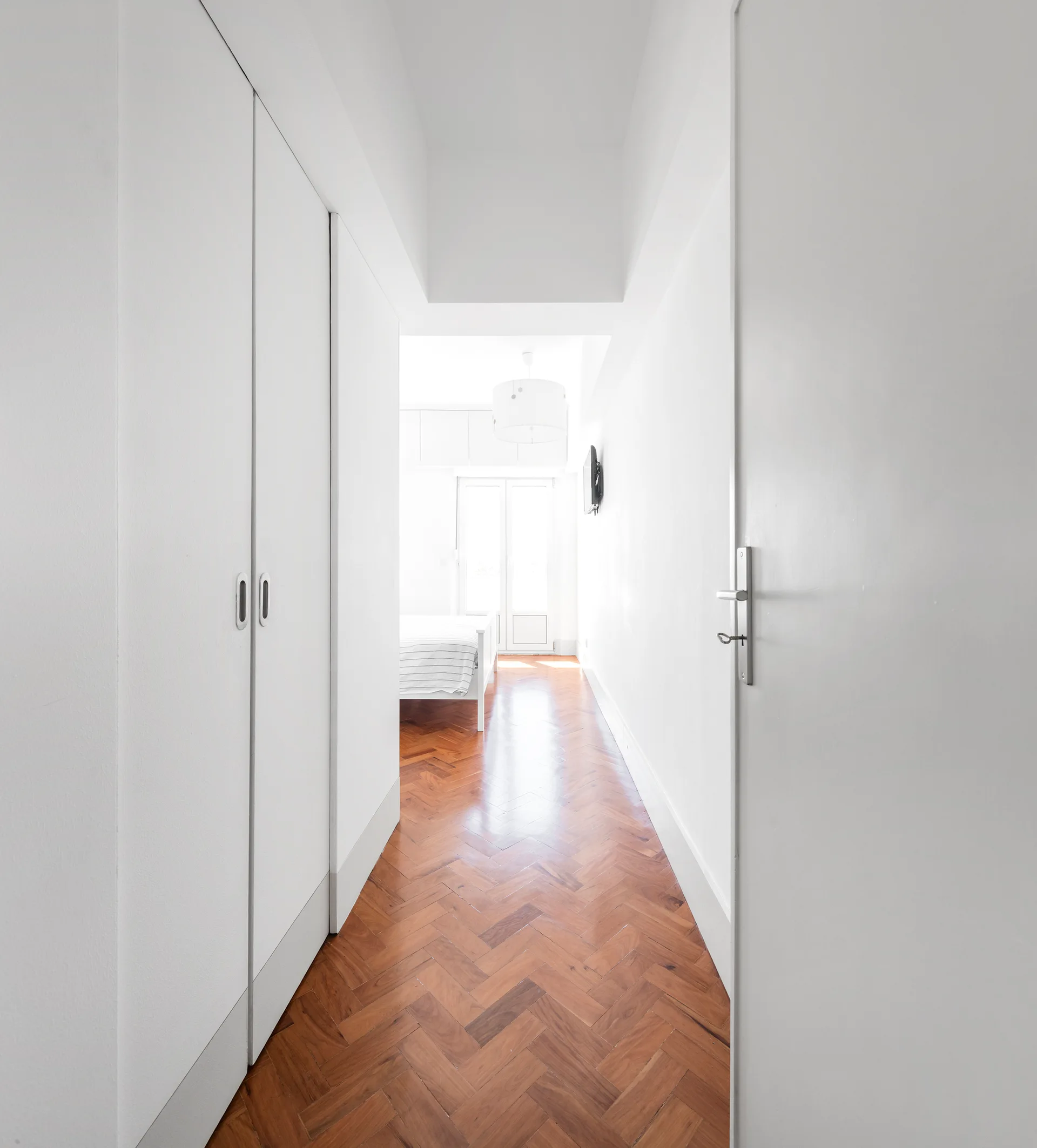A narrow white hallway features built-in white closets on one side and a warm brown herringbone wood floor leading toward a bright bedroom.