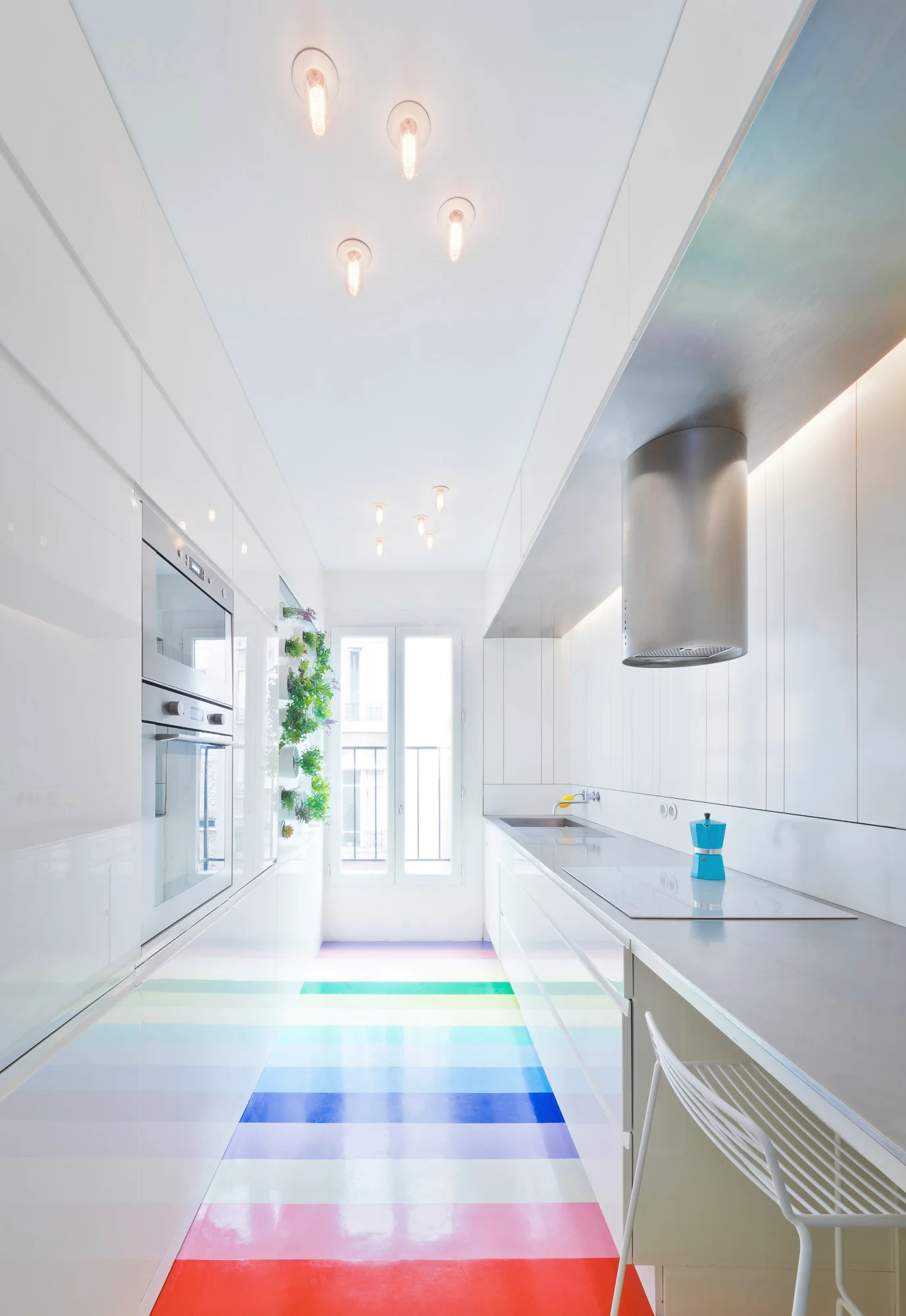 A narrow galley kitchen features high-gloss white cabinets and a glossy rainbow-striped floor leading to a window.