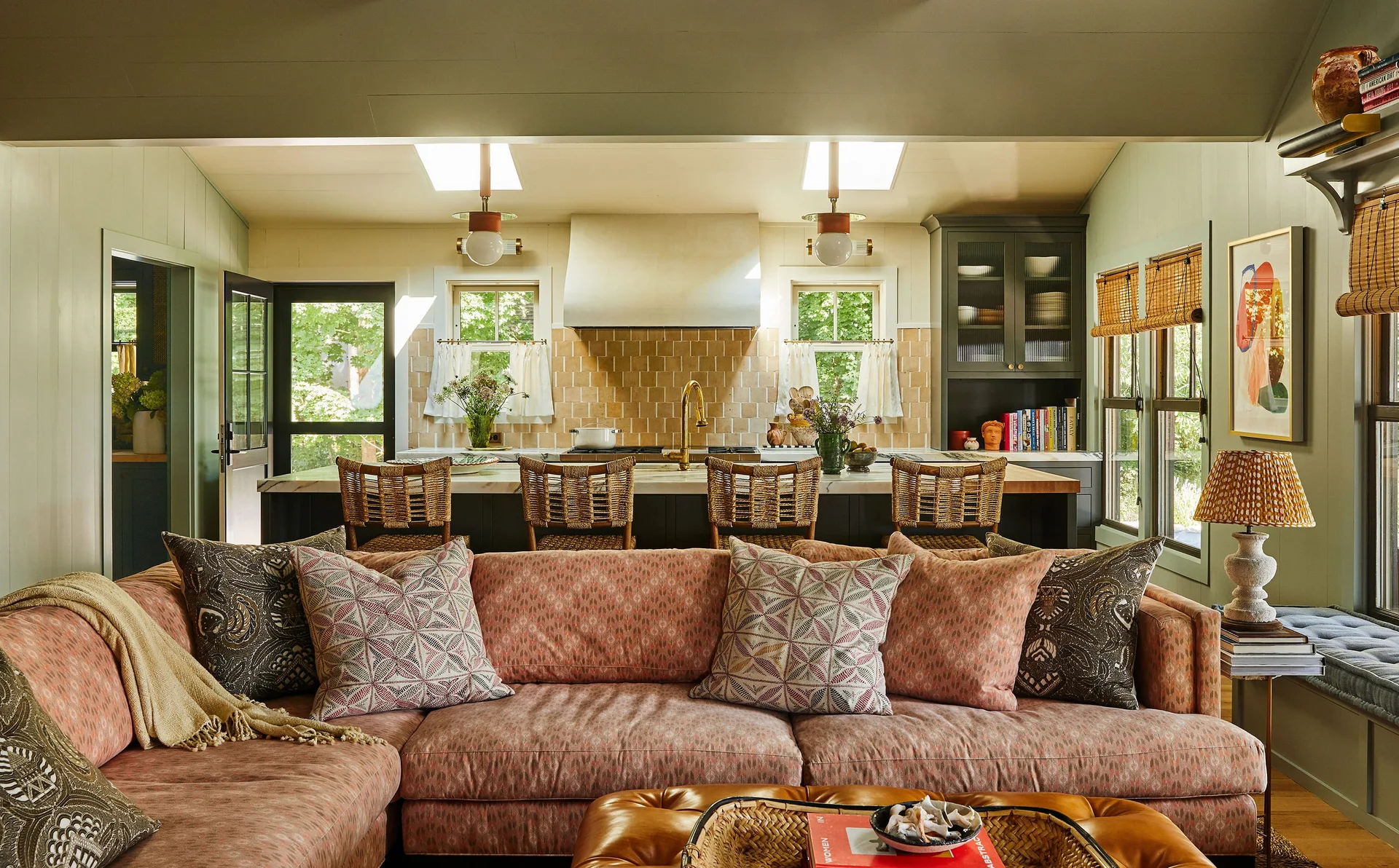 A patterned pink sectional sofa sits in front of an open kitchen featuring a marble island, woven bar stools, and sage green walls.