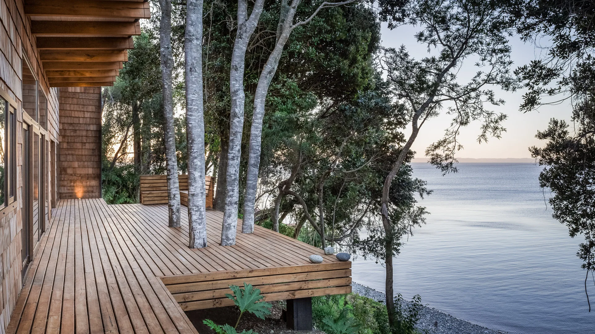 An outdoor wooden deck built around mature trees overlooks a calm waterfront view next to a house with shingle siding.