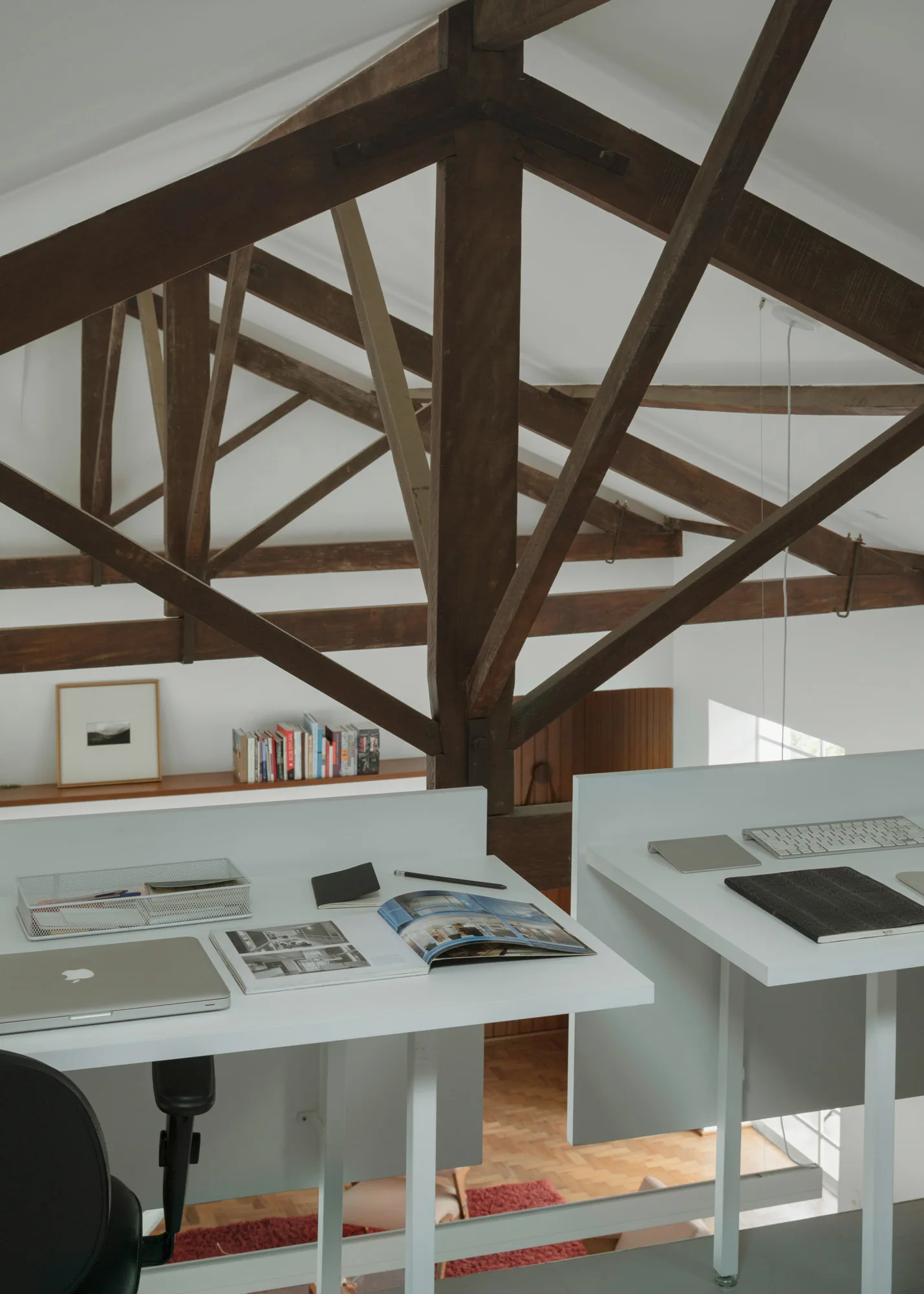 Two minimalist white desks with laptops and organizers sit under heavy exposed dark wood ceiling trusses in a loft-style home office.