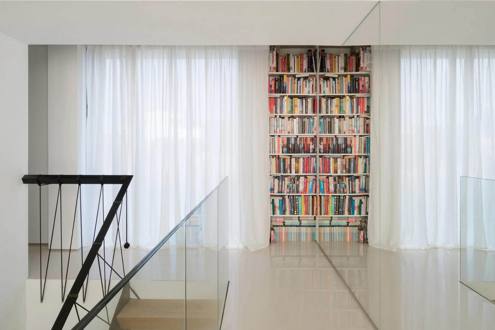 A bright white hallway featuring a built-in bookshelf next to white sheer curtains, a mirrored wall, and a staircase with a glass railing.