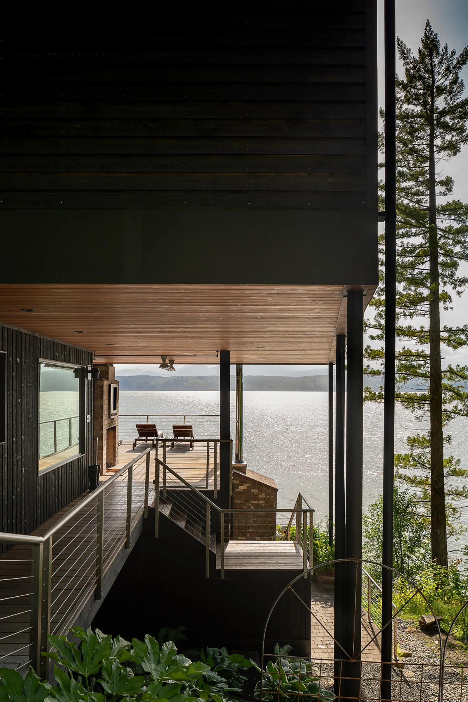 A multi-level modern wooden deck with charcoal siding, metal cable railings, and outdoor chairs overlooking a large body of water.