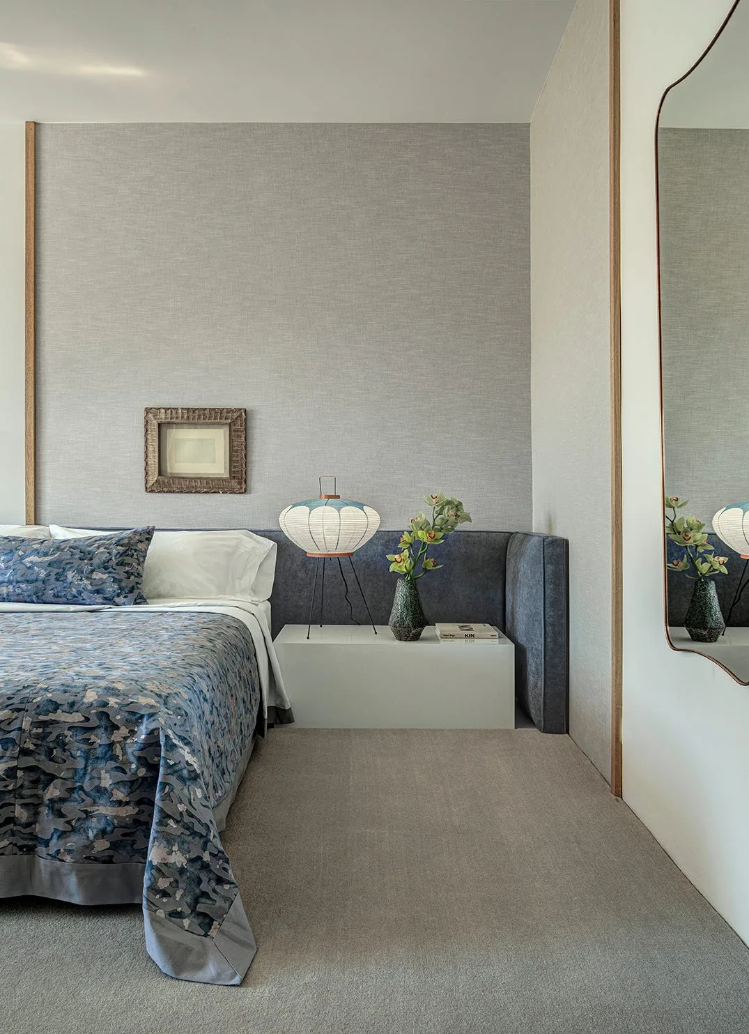 A modern bedroom with textured grey wallpaper features a blue patterned bed, a low white nightstand, and a tall wavy-edged wall mirror.