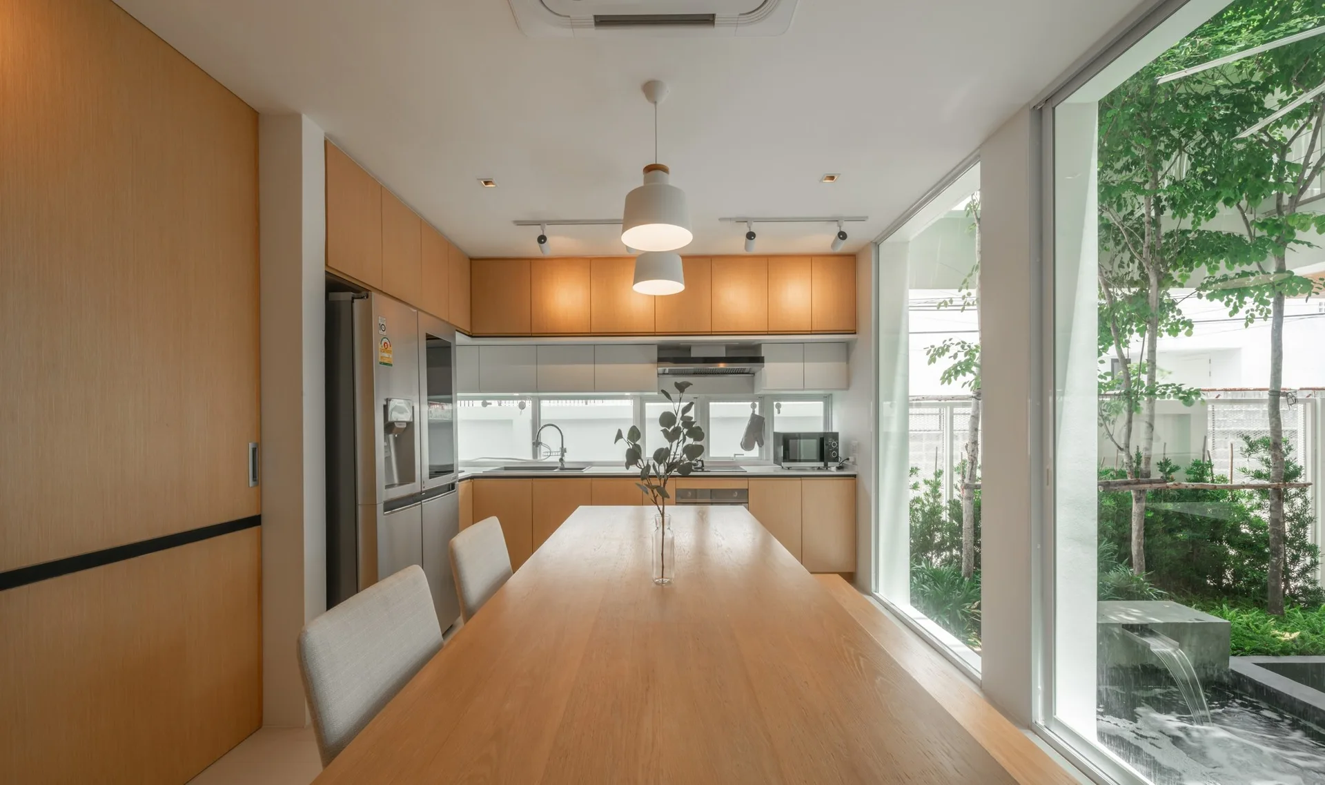 A light wood kitchen and dining area with a long table next to a floor-to-ceiling glass window showing an outdoor garden and pond.