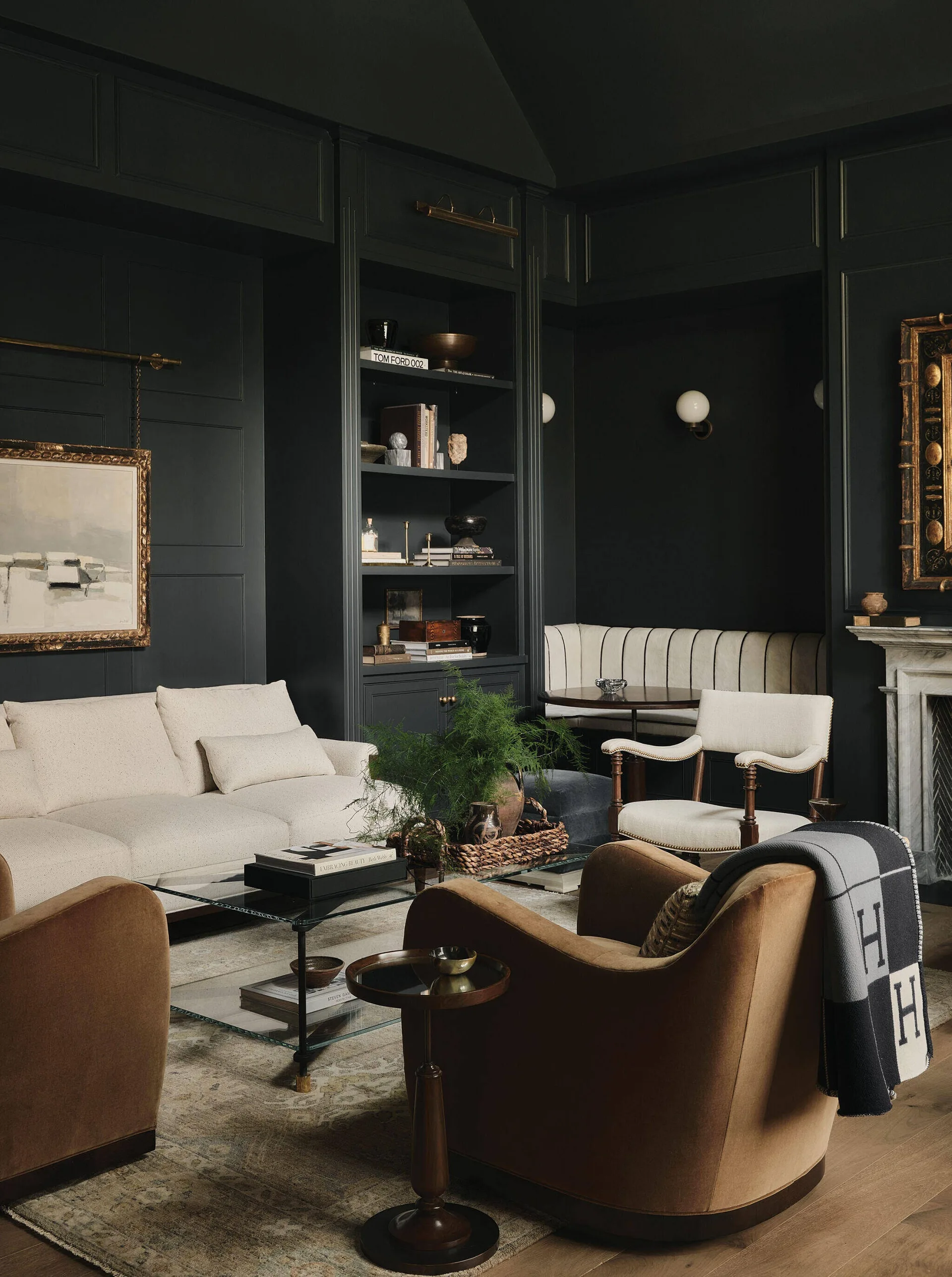 A moody living room features dark charcoal paneled walls, a cream sofa, brown velvet armchairs, and built-in bookshelves.