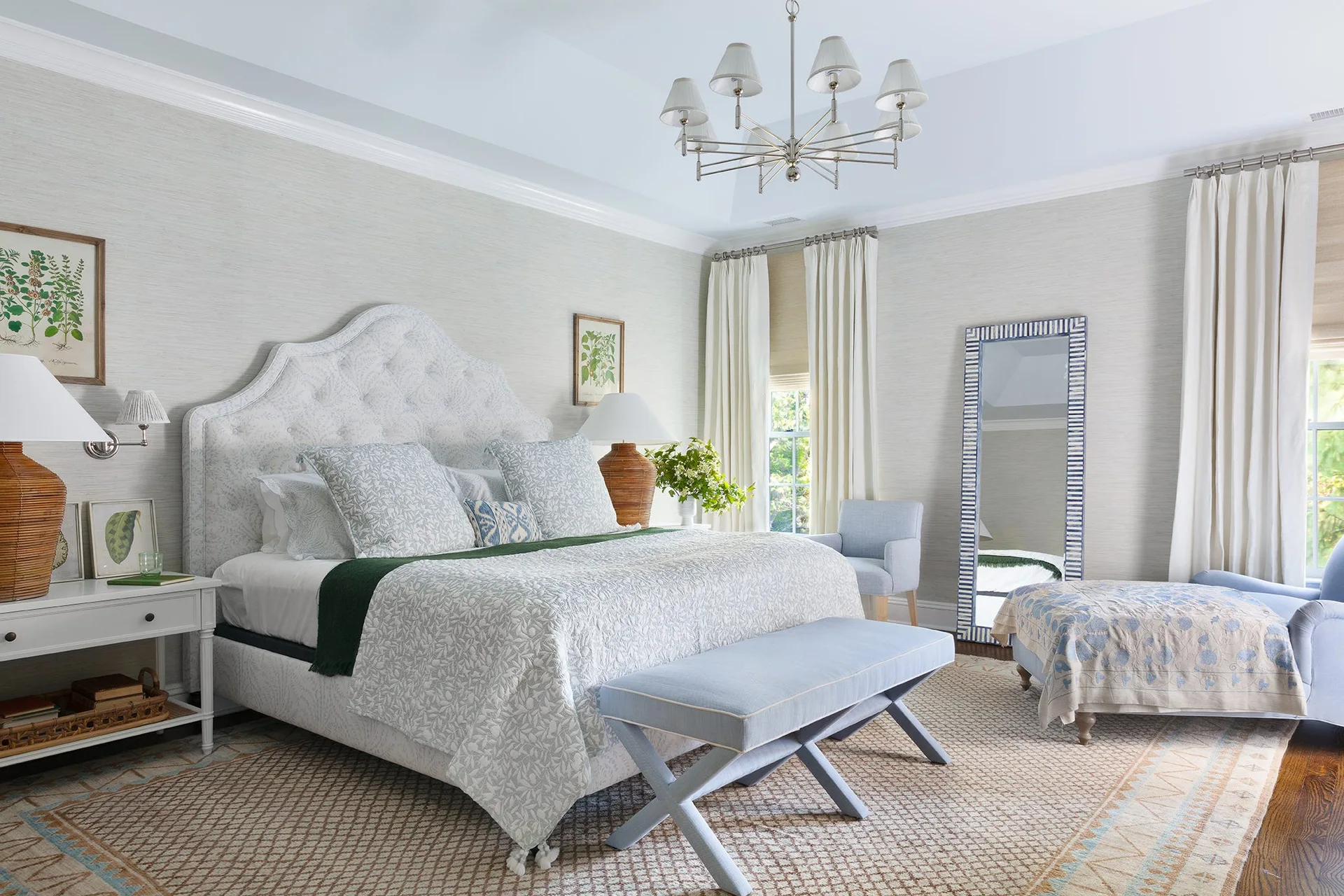 A bedroom features a patterned tufted headboard, a blue upholstered bench, woven table lamps, textured wallpaper, and a large floor mirror.