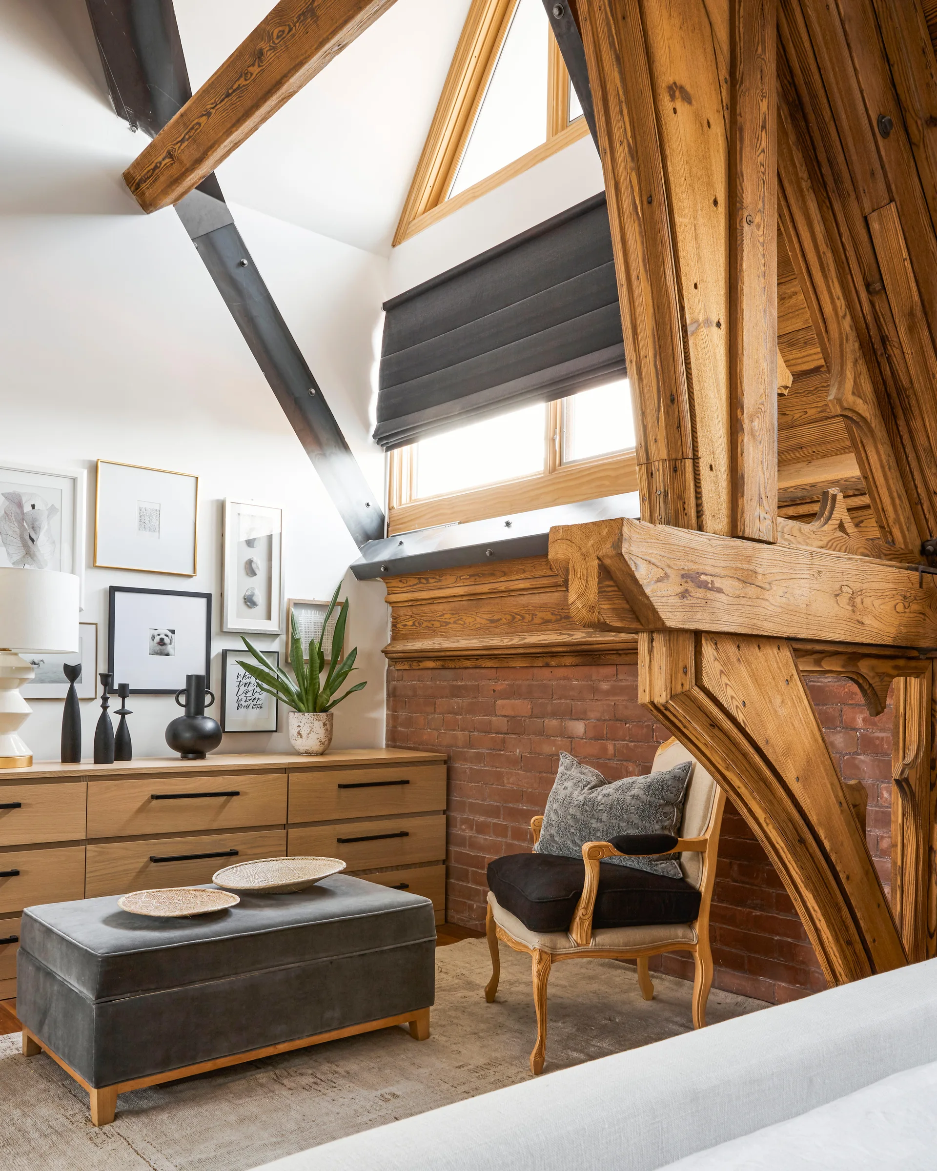 A loft bedroom features heavy wood ceiling beams, an exposed red brick wall, a light wood dresser with framed art, and a grey upholstered ottoman.