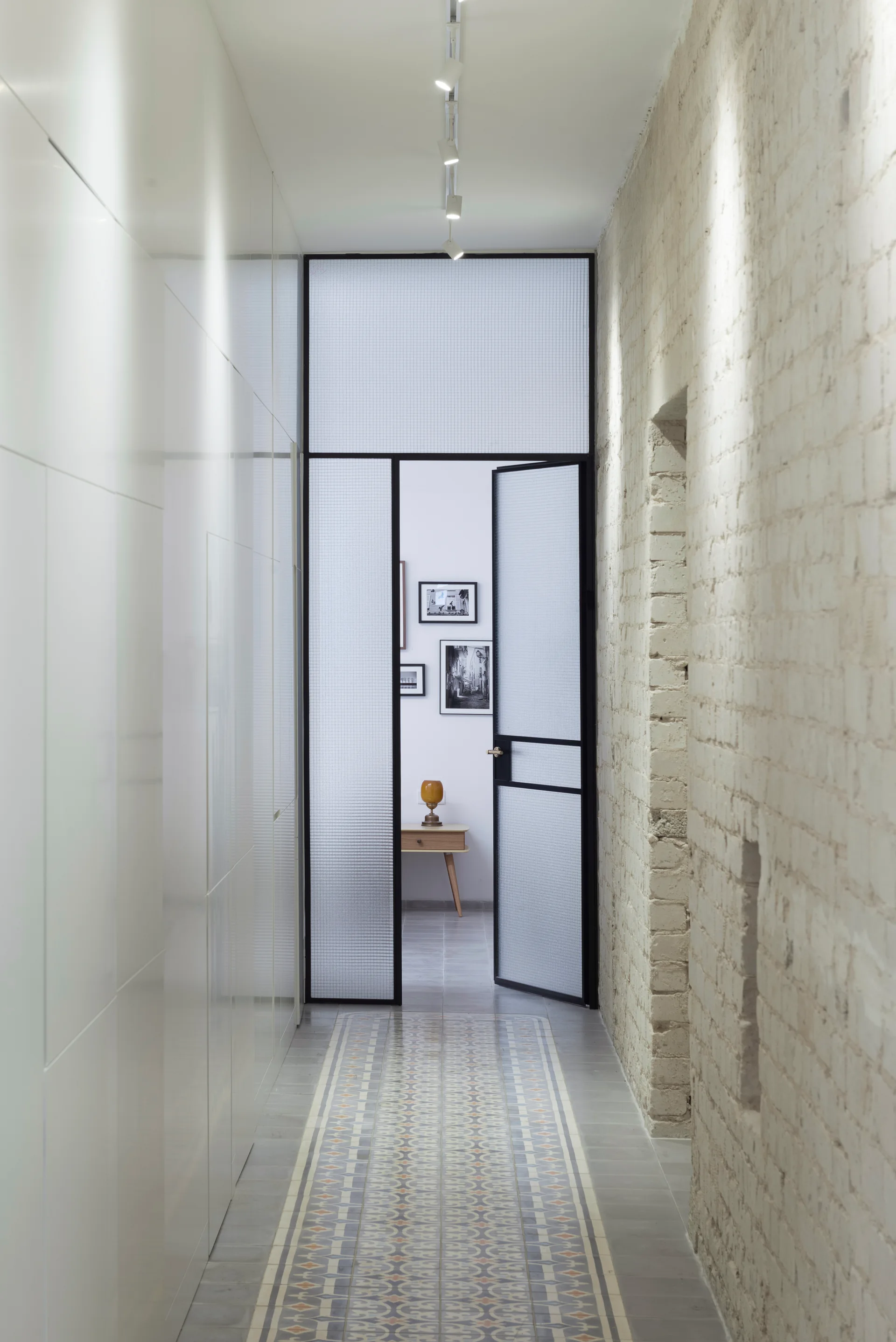 A narrow hallway featuring a textured white brick wall, glossy floor-to-ceiling white cabinets, a patterned tile runner, and a black-framed glass door.