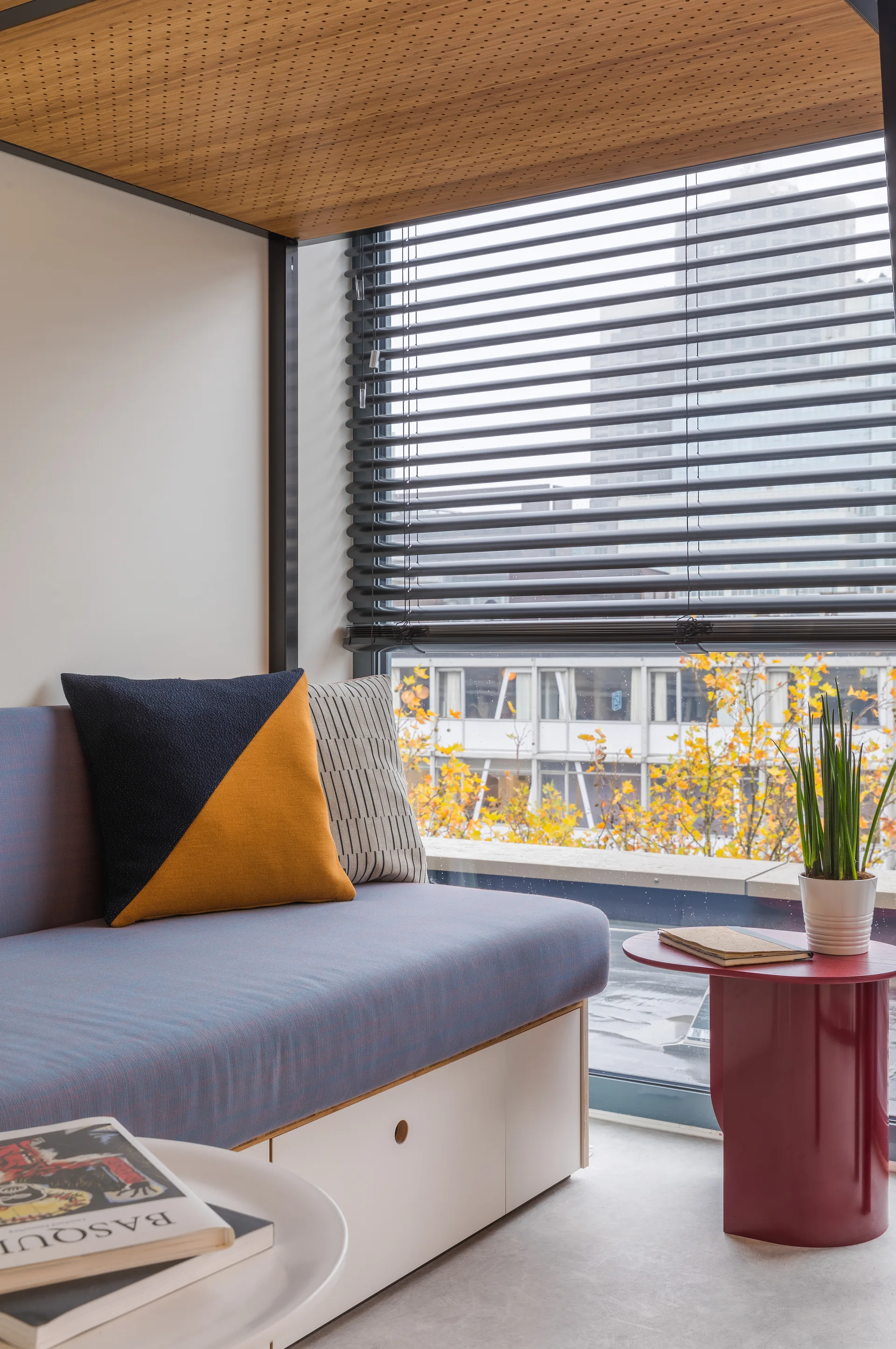 A modern built-in daybed with blue cushions and geometric pillows sits in a window alcove below a perforated wood ceiling panel.