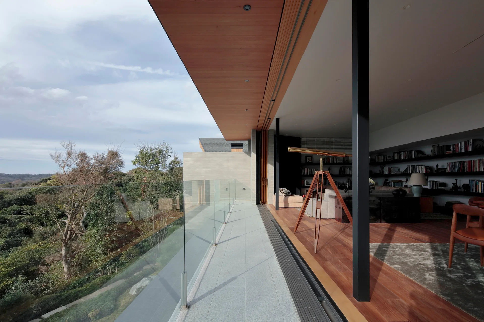 A modern stone terrace with a frameless glass railing overlooks trees, transitioning into an indoor living area with wood flooring and a telescope.