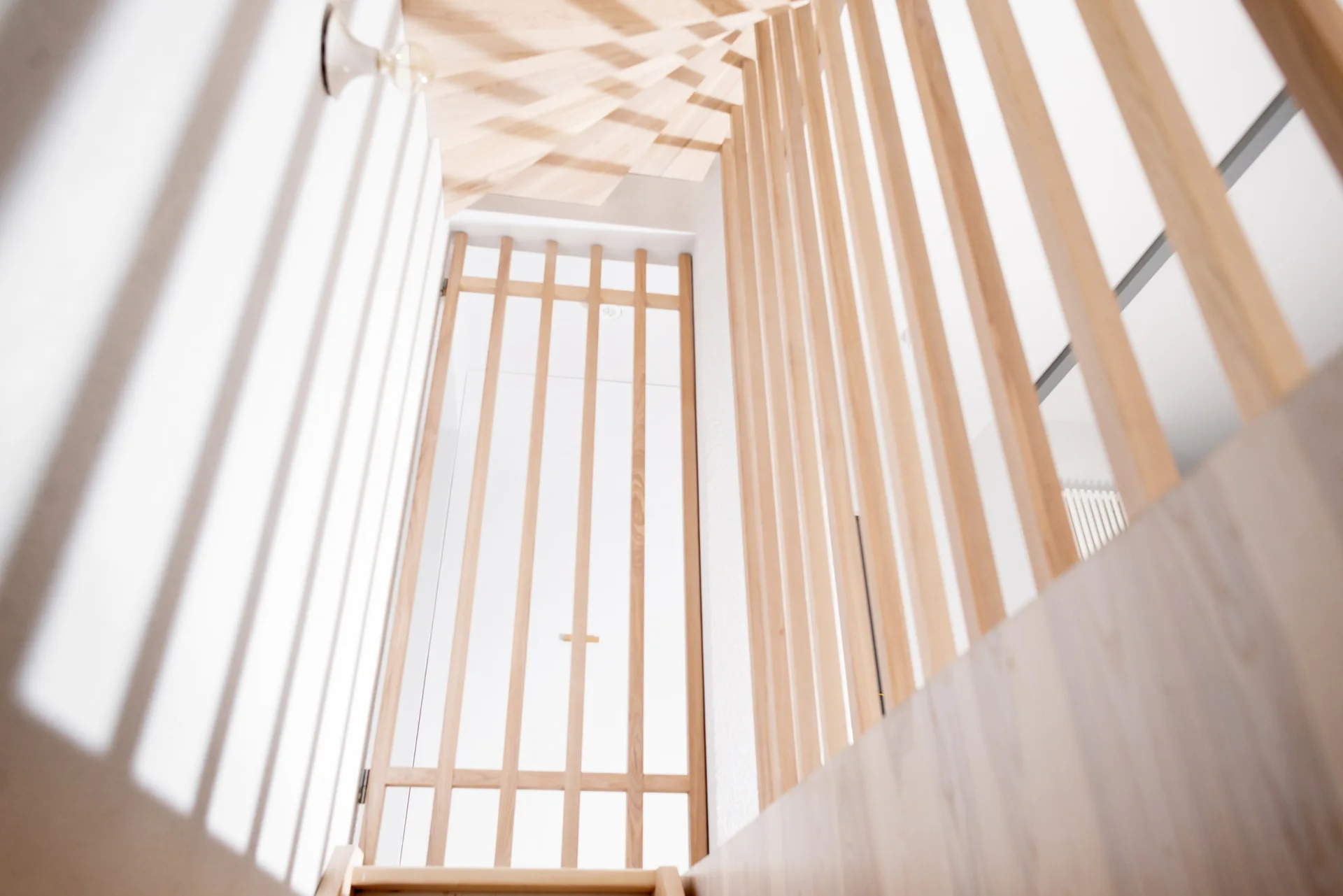 A vertical view looking up a stairwell with a light wood vertical slat screen railing casting shadows on white walls.
