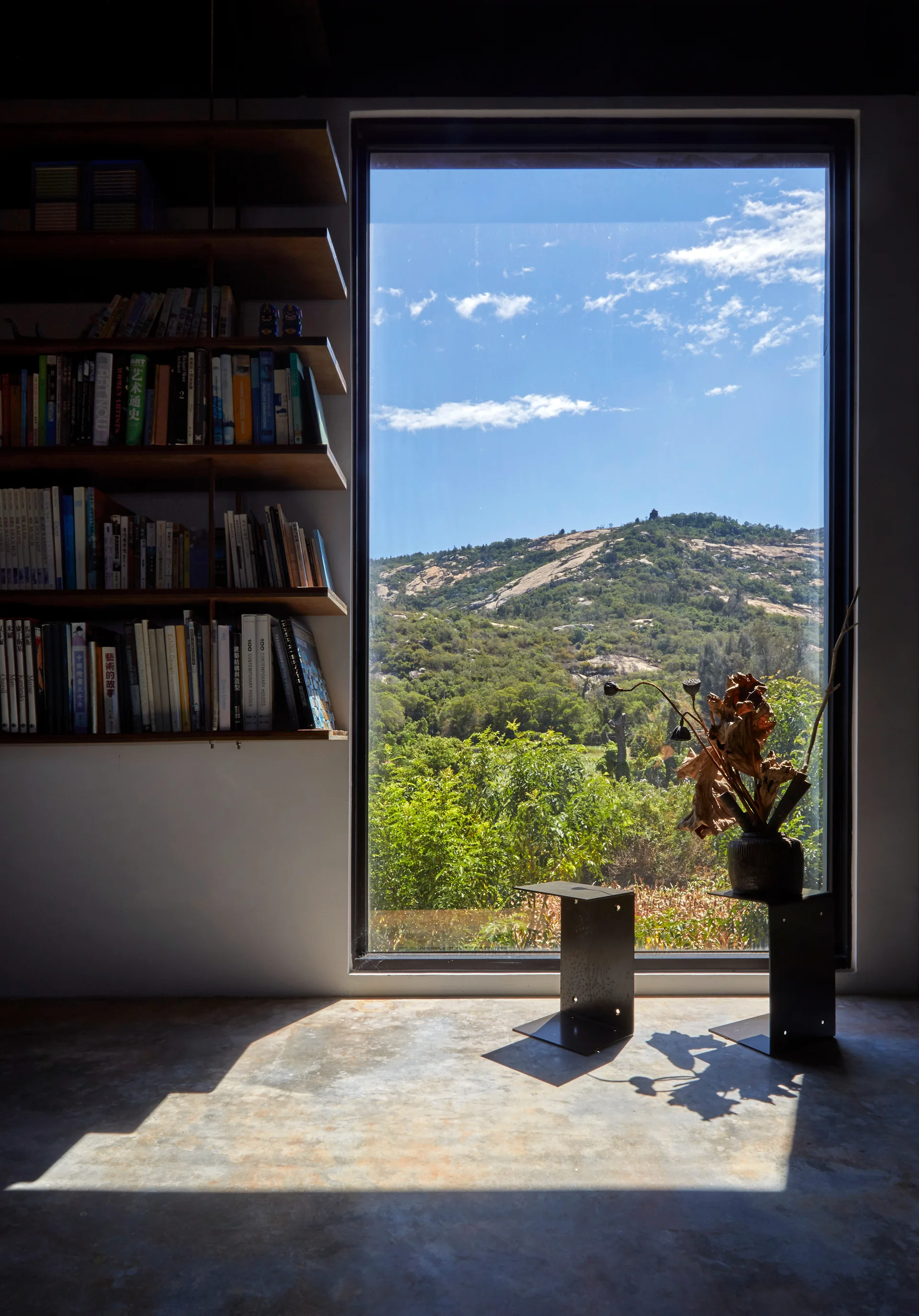 A minimalist library corner features a large picture window framing a mountain view, a wooden bookshelf, and small metal tables on a concrete floor.
