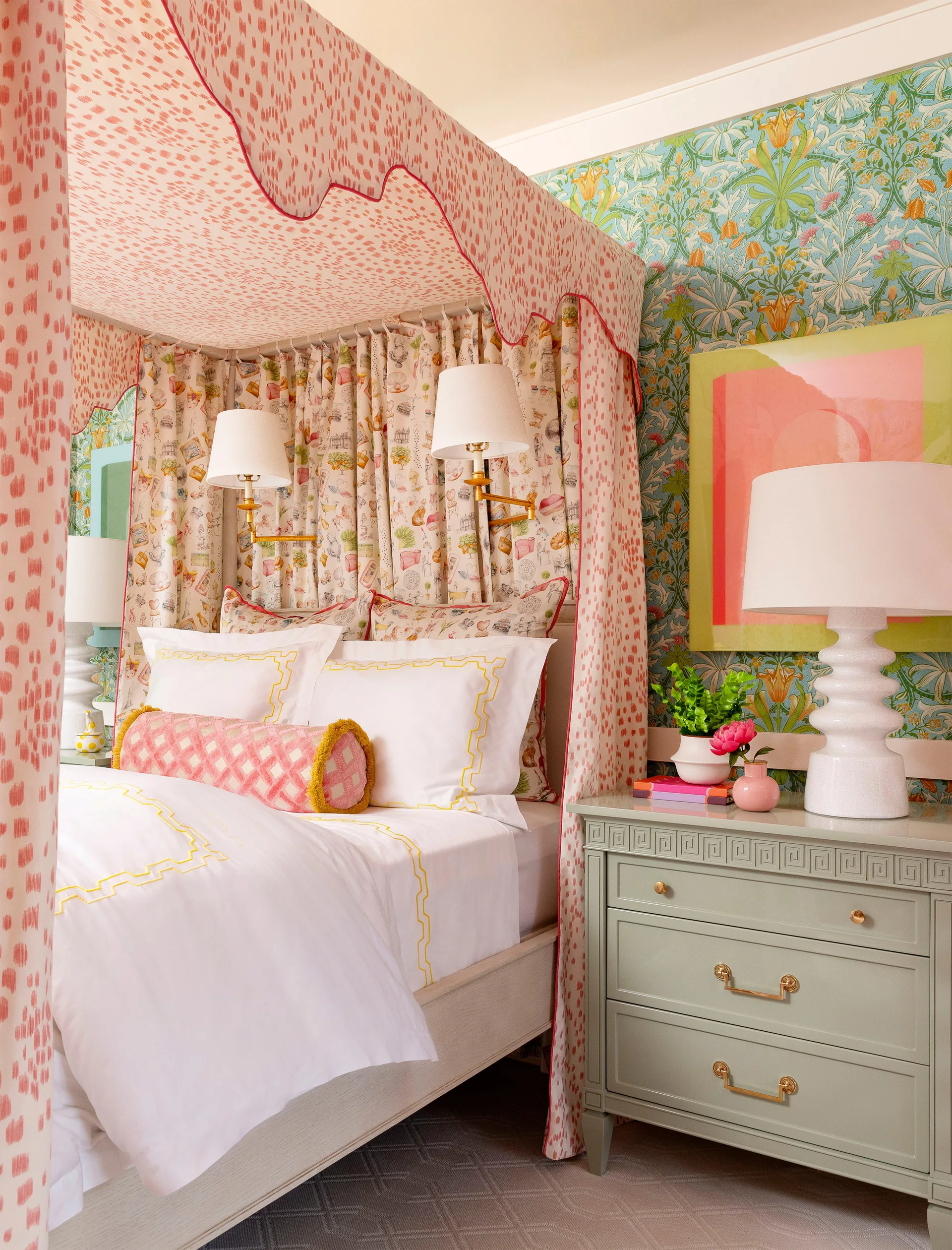 A bedroom features a pink patterned canopy bed with a scalloped valance next to a sage green nightstand against green floral wallpaper.