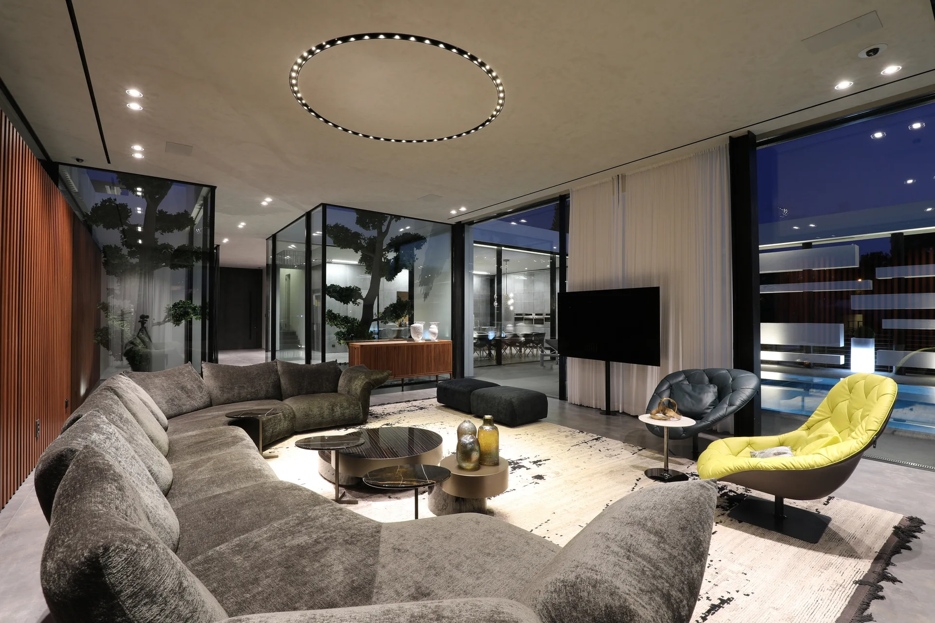 A modern living room features a large curved grey sectional sofa, a bright yellow armchair, and a circular ceiling ring light.