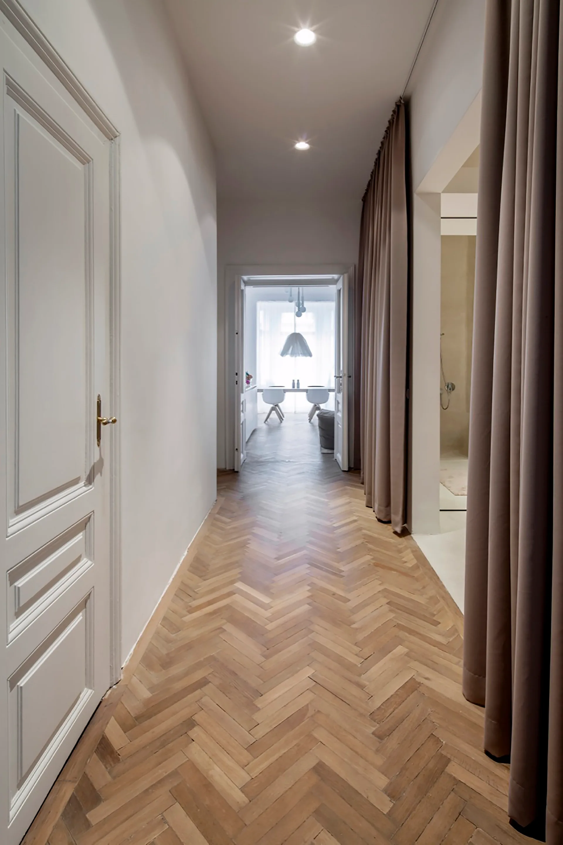 A residential hallway features herringbone wood flooring, a white paneled door, and long taupe curtains leading toward an open doorway.