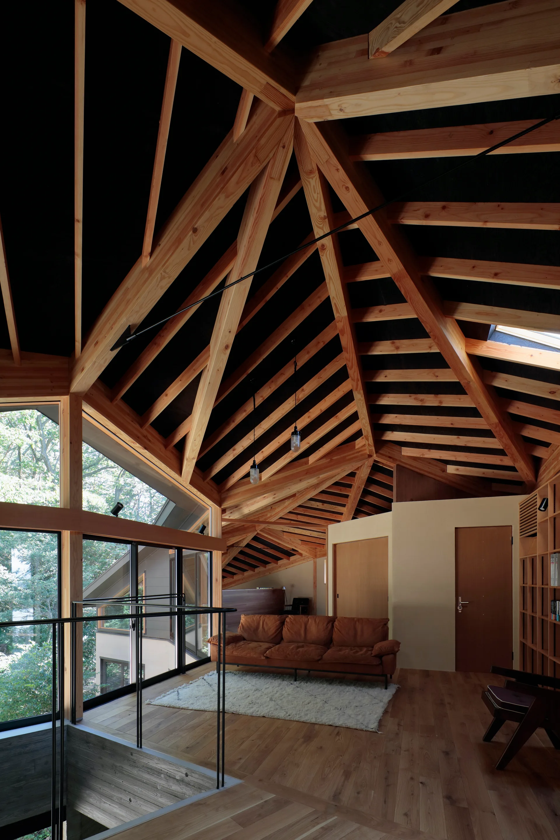 A modern loft living room features a dramatic exposed timber vaulted ceiling, a brown leather sofa, and large windows looking out to trees.