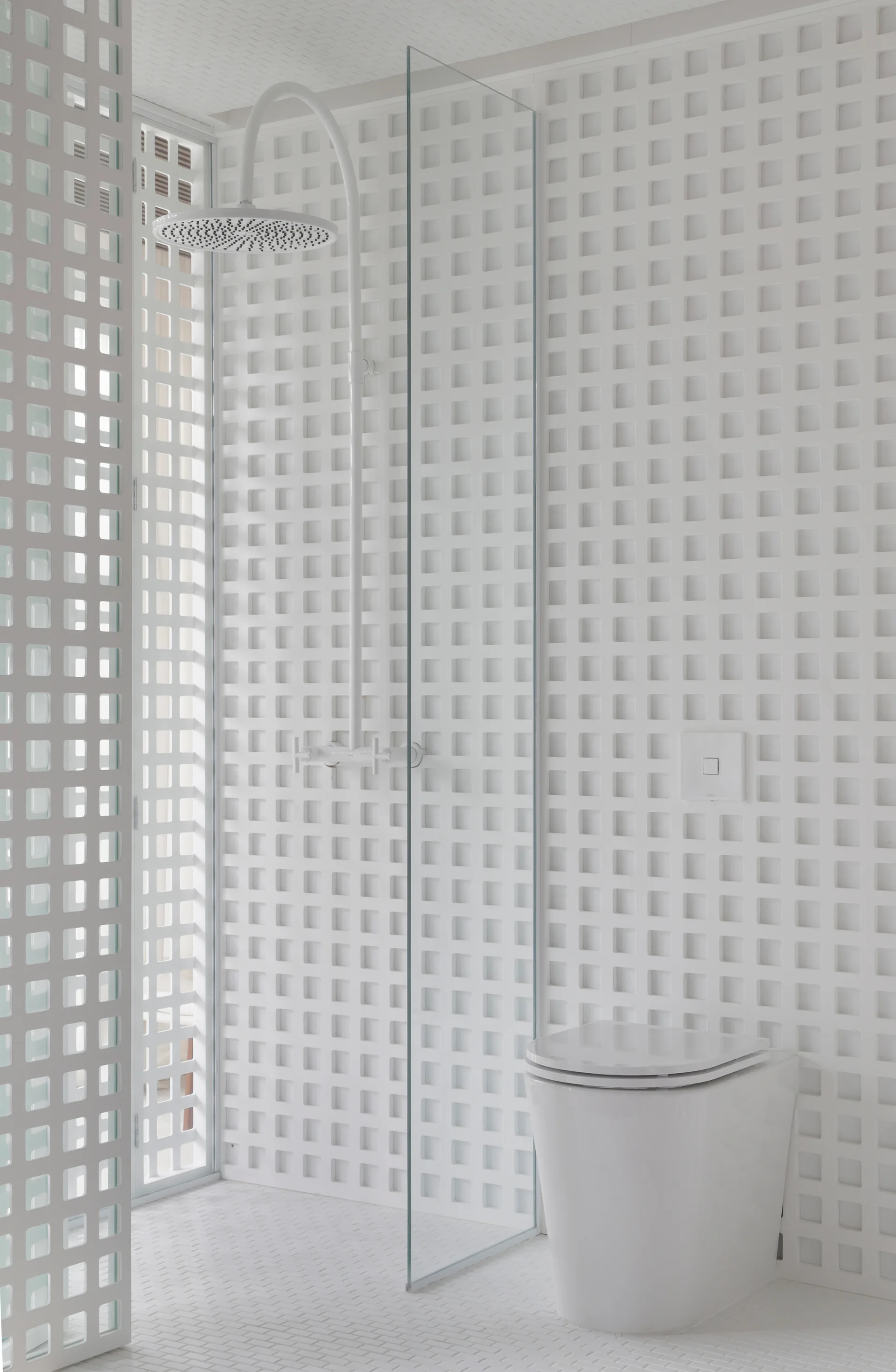An all-white bathroom features a prominent square grid pattern on the walls, a frameless glass shower panel, and a white toilet.