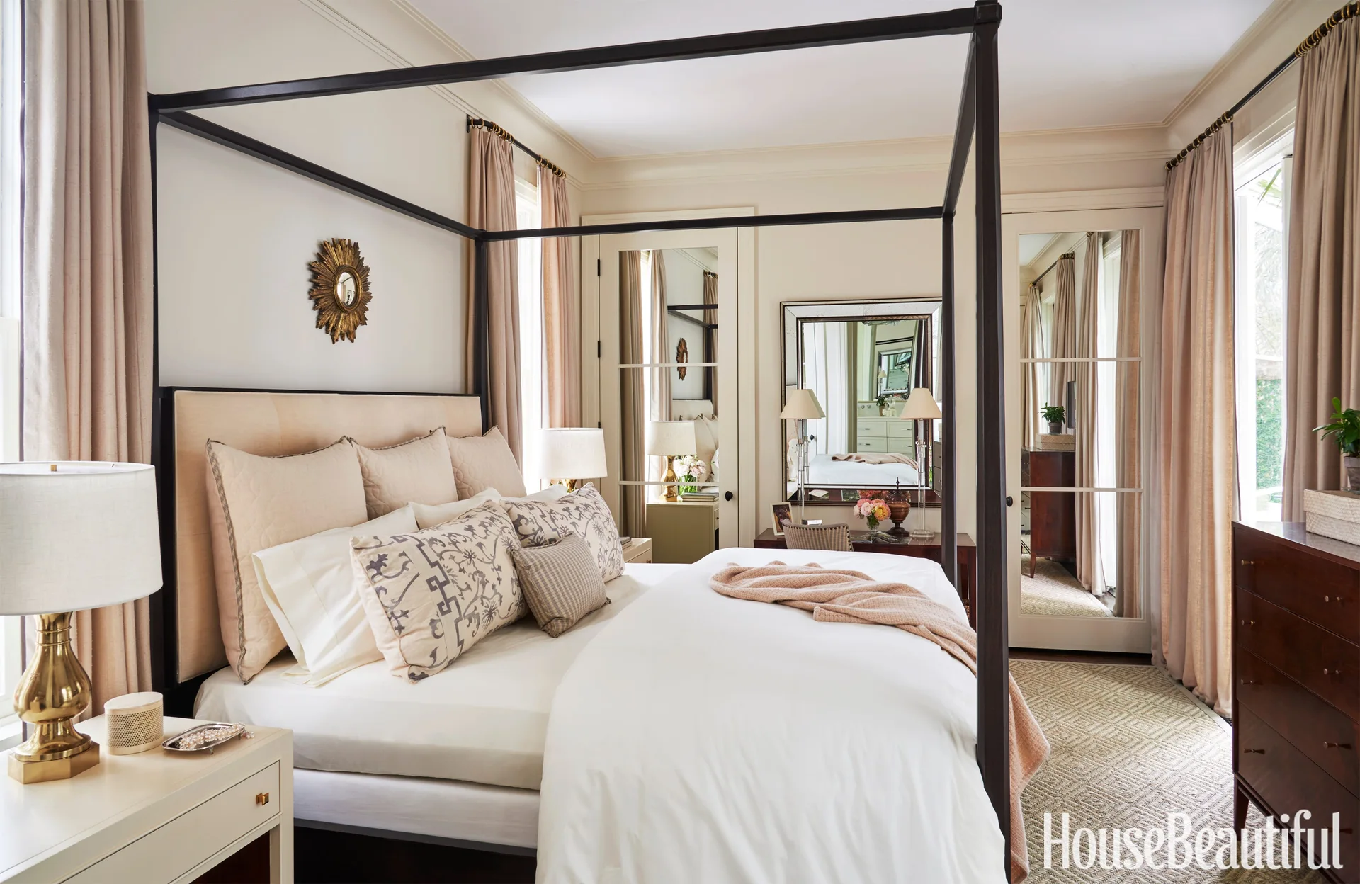 A neutral bedroom features a black metal canopy bed, beige curtains, brass table lamps, and mirrored double doors reflecting the room.