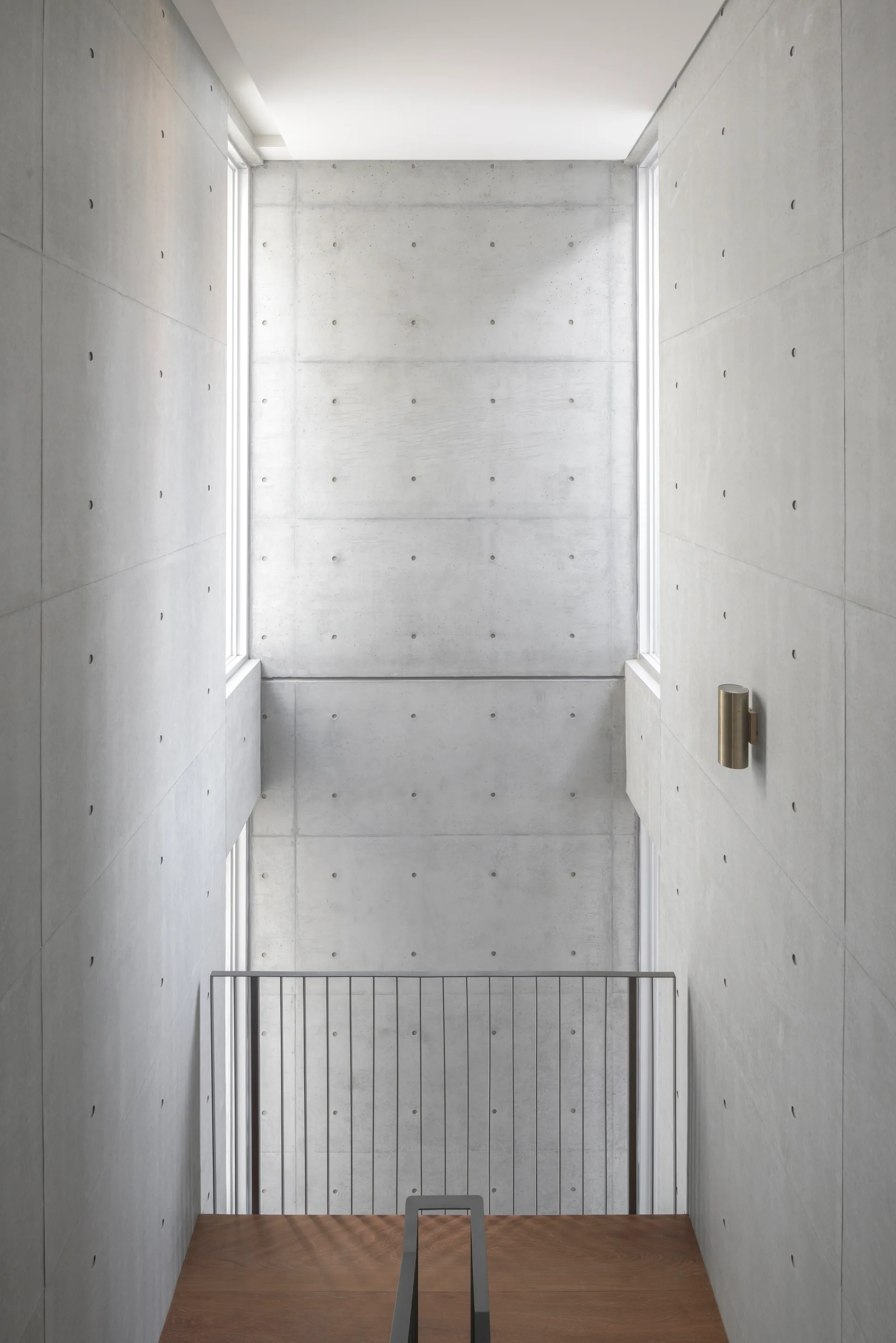 A modern staircase features warm wooden steps and a black metal railing set against tall, raw exposed concrete walls with vertical slot windows.