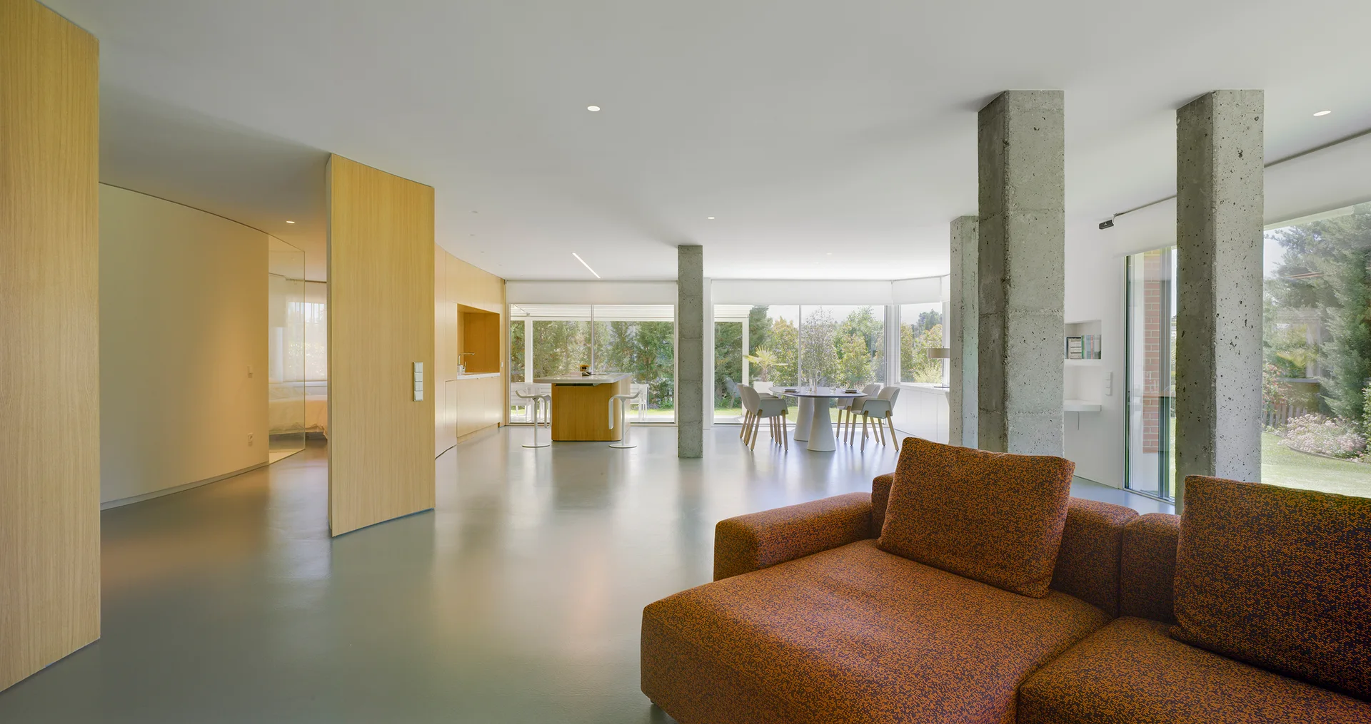 An open floor plan living room features an orange textured sectional sofa, exposed concrete pillars, and light wood wall paneling.