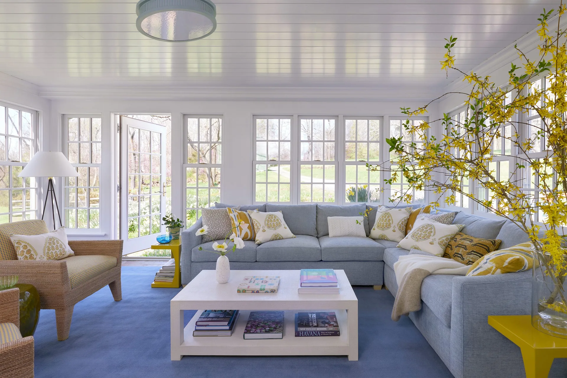 A bright sunroom featuring a light blue sectional sofa, a white coffee table, and a wicker armchair on a blue rug surrounded by grid windows.