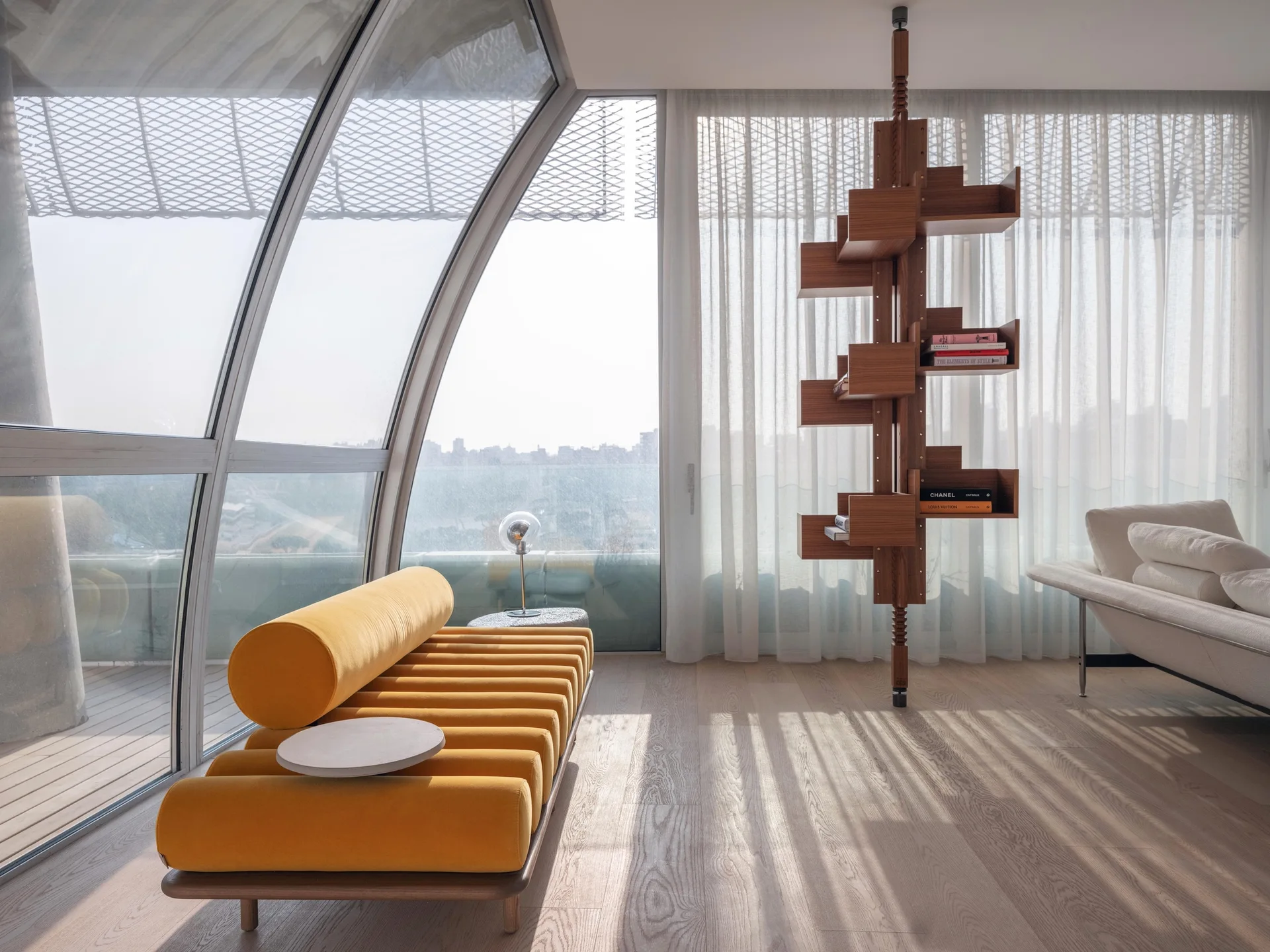 A sunlit modernist interior featuring a bright yellow daybed, a curved glass wall, and a vertical wood pole bookshelf.