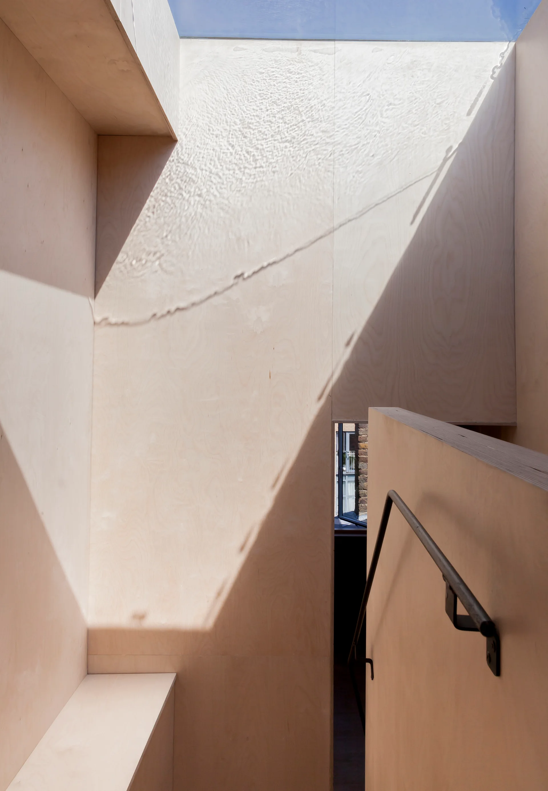 A minimalist interior staircase built with light plywood panels features a dark metal handrail and a bright rectangular skylight above.