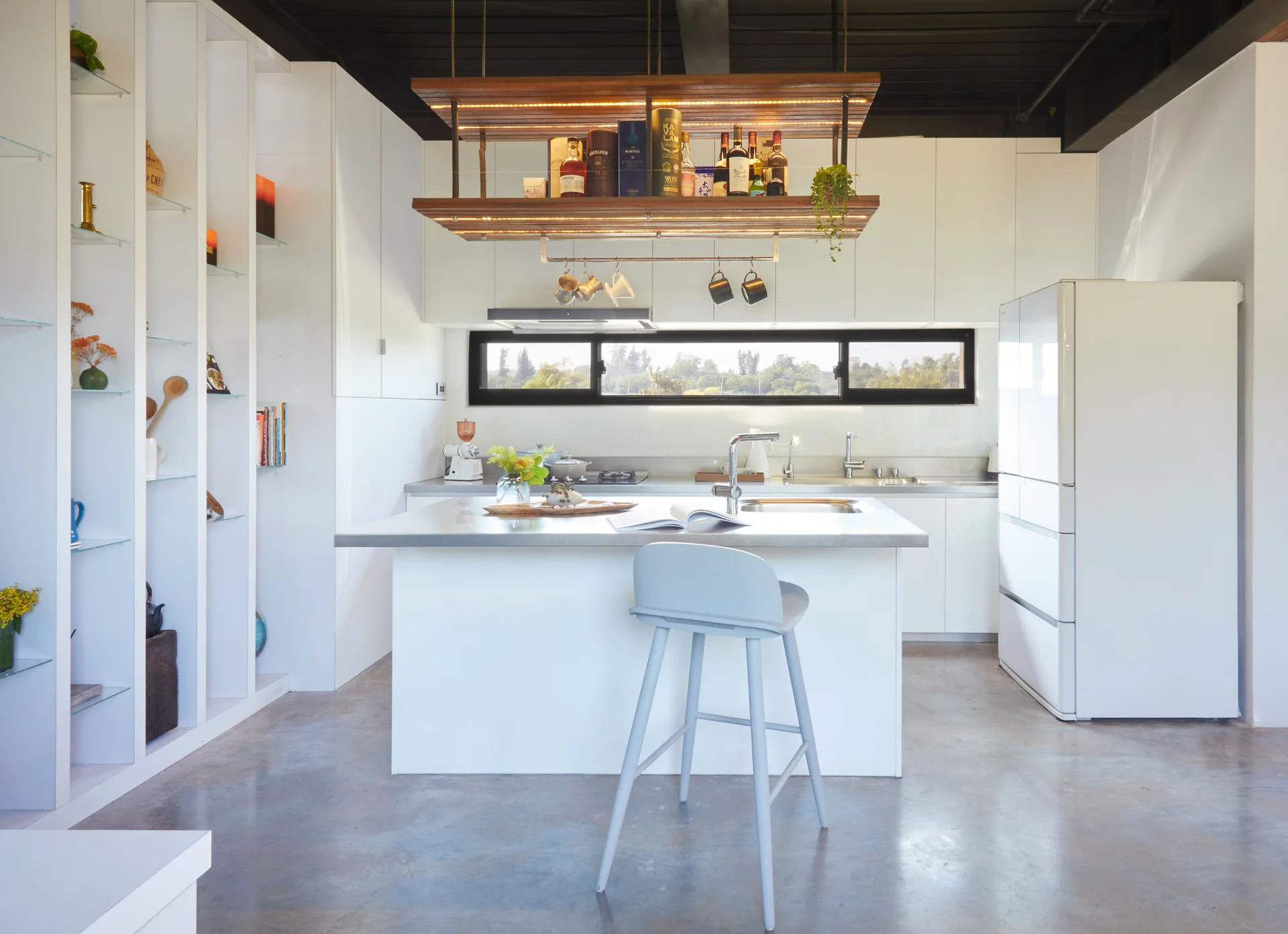 A white minimalist kitchen featuring a central island, a grey bar stool, a suspended wooden shelf, and polished concrete flooring.