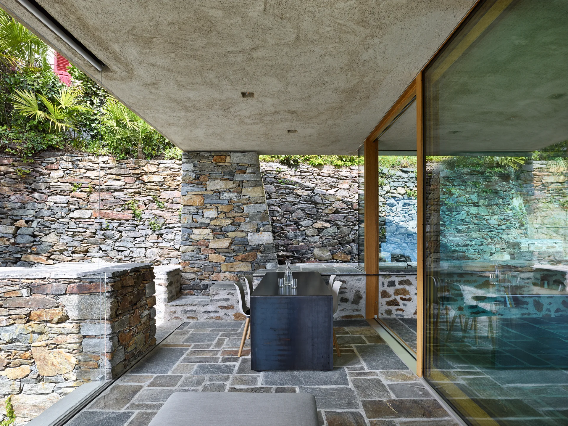 A covered outdoor dining patio features a dark rectangular table on flagstone flooring next to stacked stone walls and a glass facade.