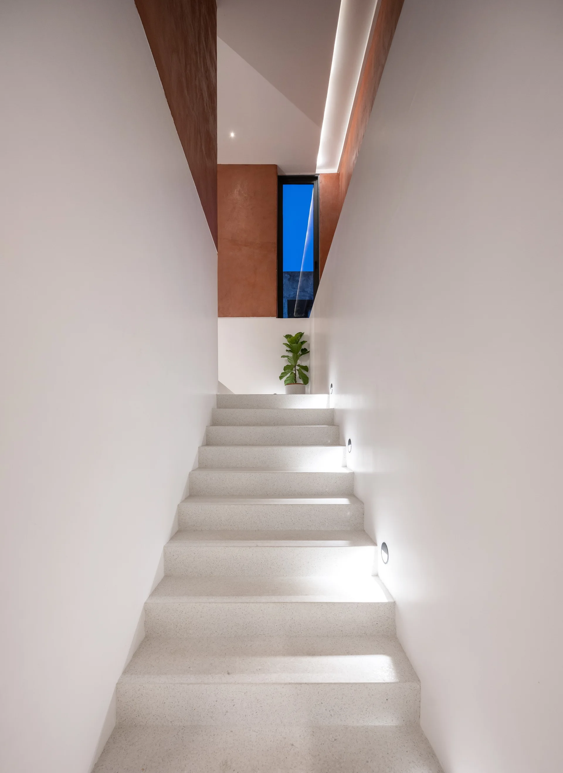 A minimalist indoor staircase featuring white terrazzo steps, recessed circular wall lights, and terracotta wall accents at the top.