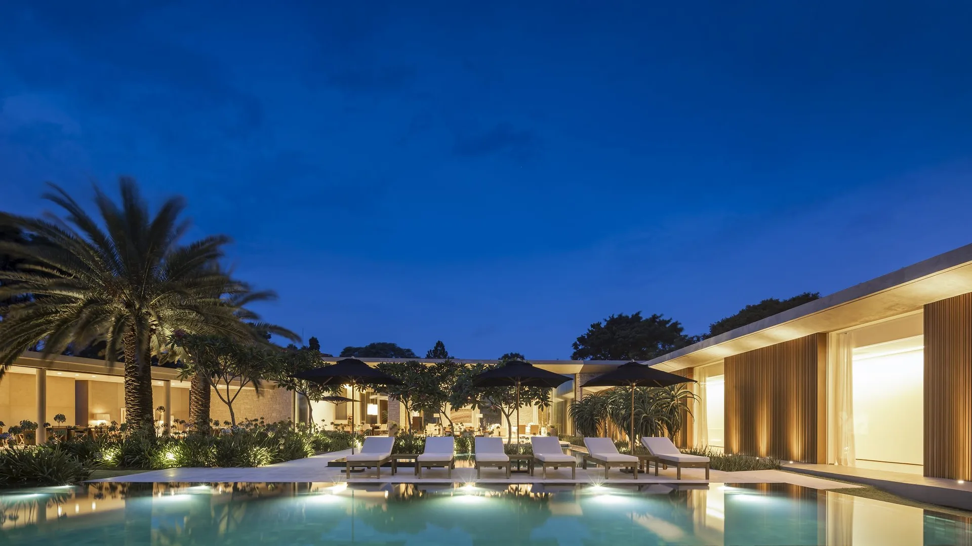 A contemporary swimming pool and deck illuminated at night with a row of lounge chairs, black umbrellas, and warm architectural uplighting.